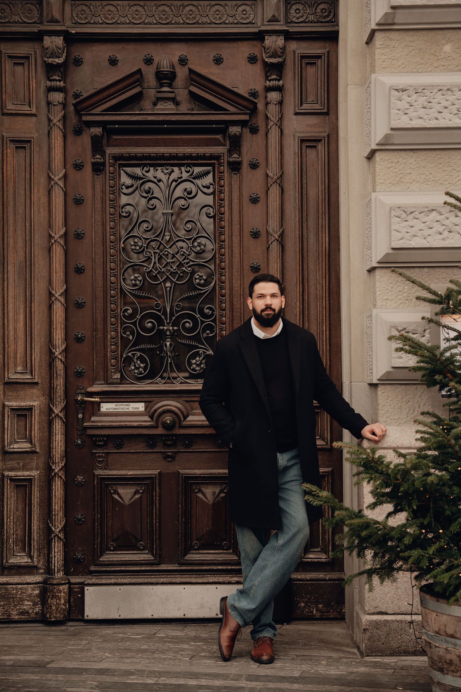 Bearded man in black coat posing against decorative carved wooden door with ironwork beside evergreen tree.