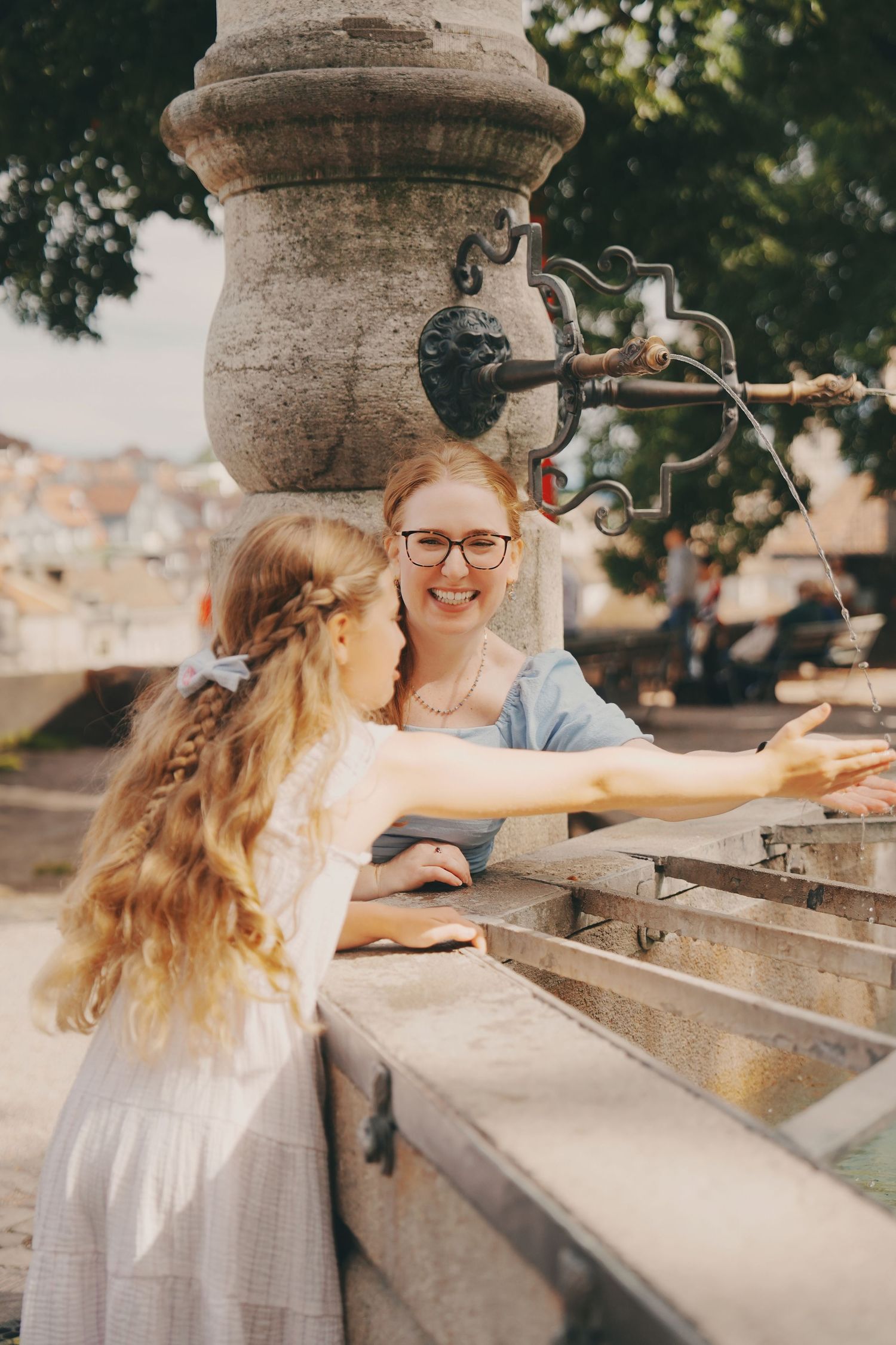 A woman with glasses and a girl in a white dress play at an ornate stone fountain outdoors.