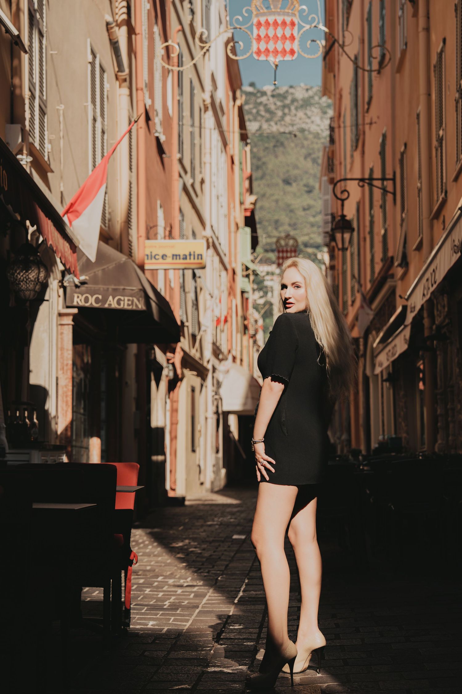 Blonde woman in black mini dress and heels posing in a European old town street.