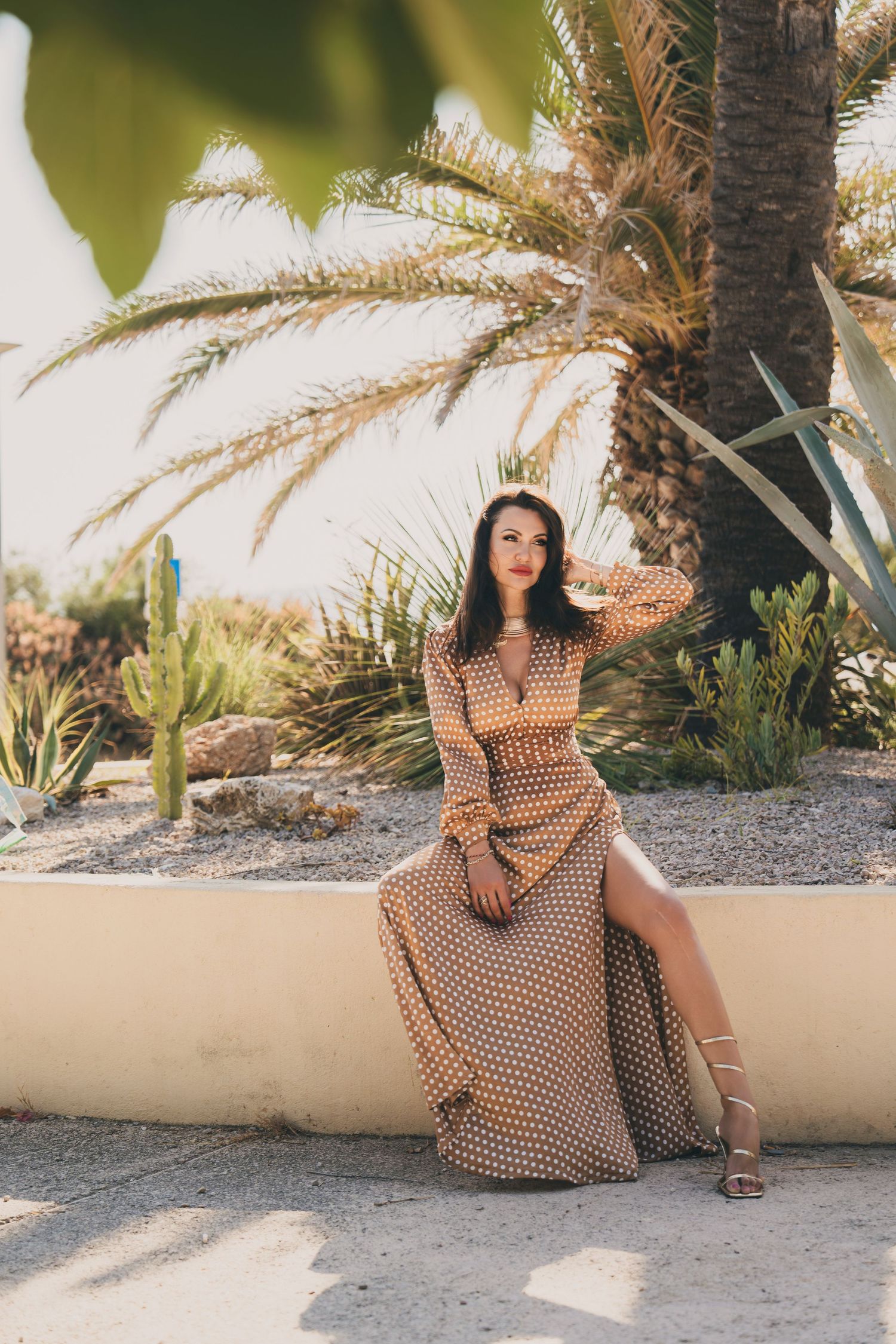Brunette woman in tan polka dot dress sitting on garden wall with tropical palm trees backdrop.
