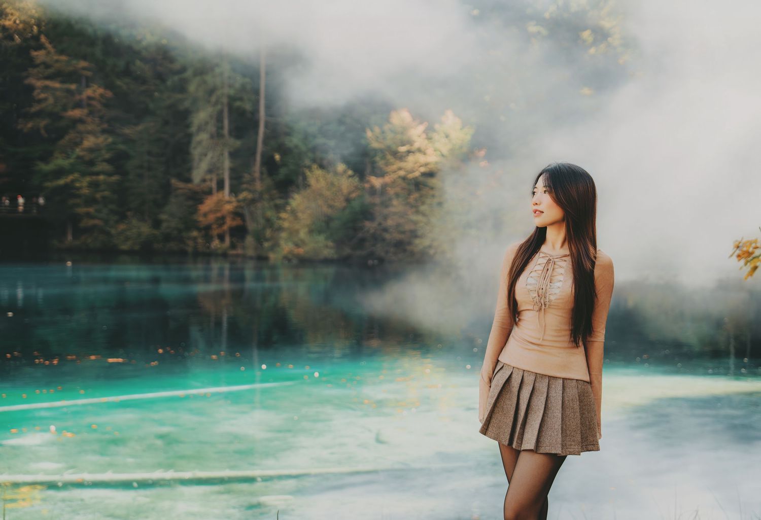 Young woman in beige outfit standing by a misty turquoise lake surrounded by autumn foliage.