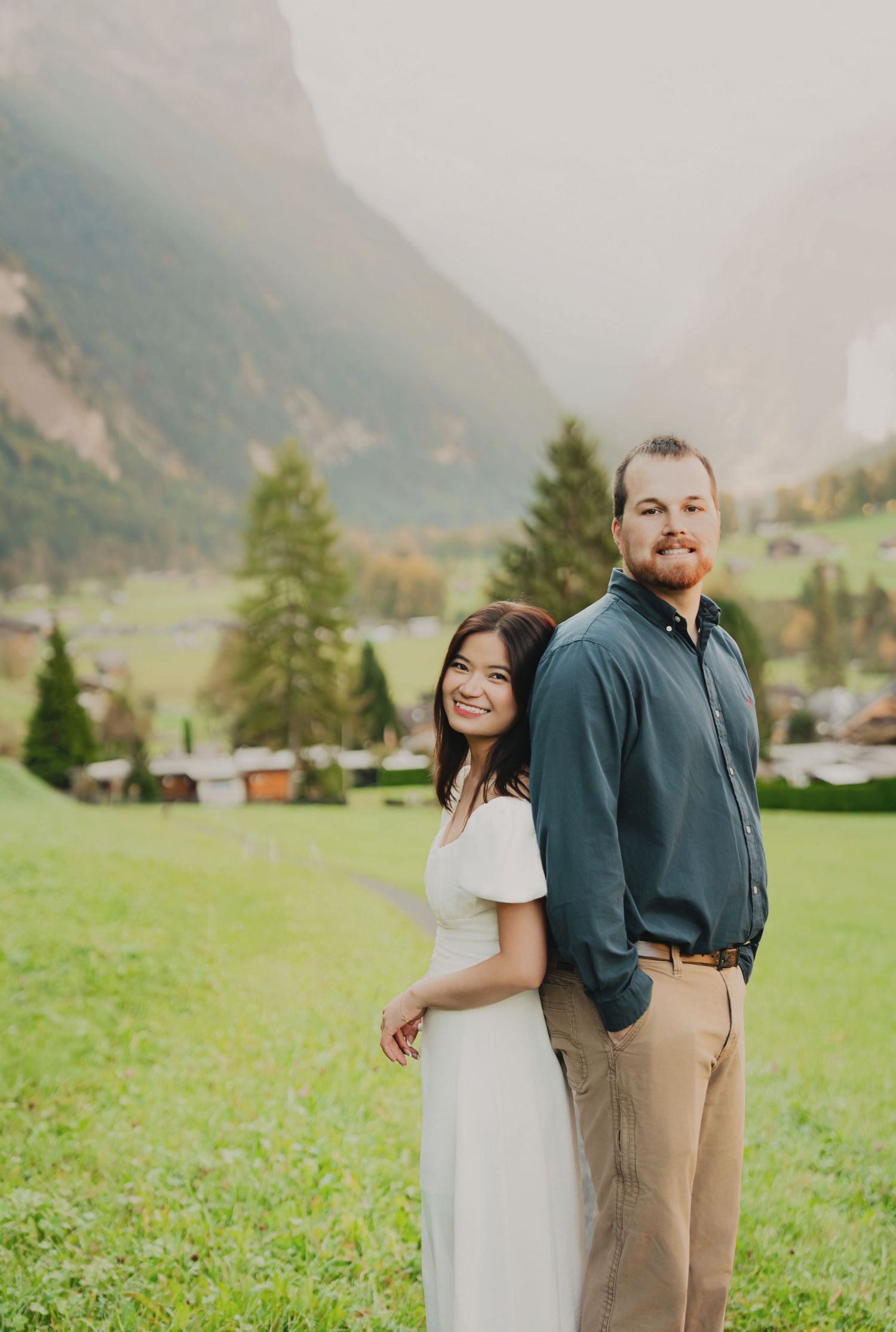 Happy couple posing back-to-back on a green meadow with a foggy alpine valley and mountain backdrop in Switzerland.