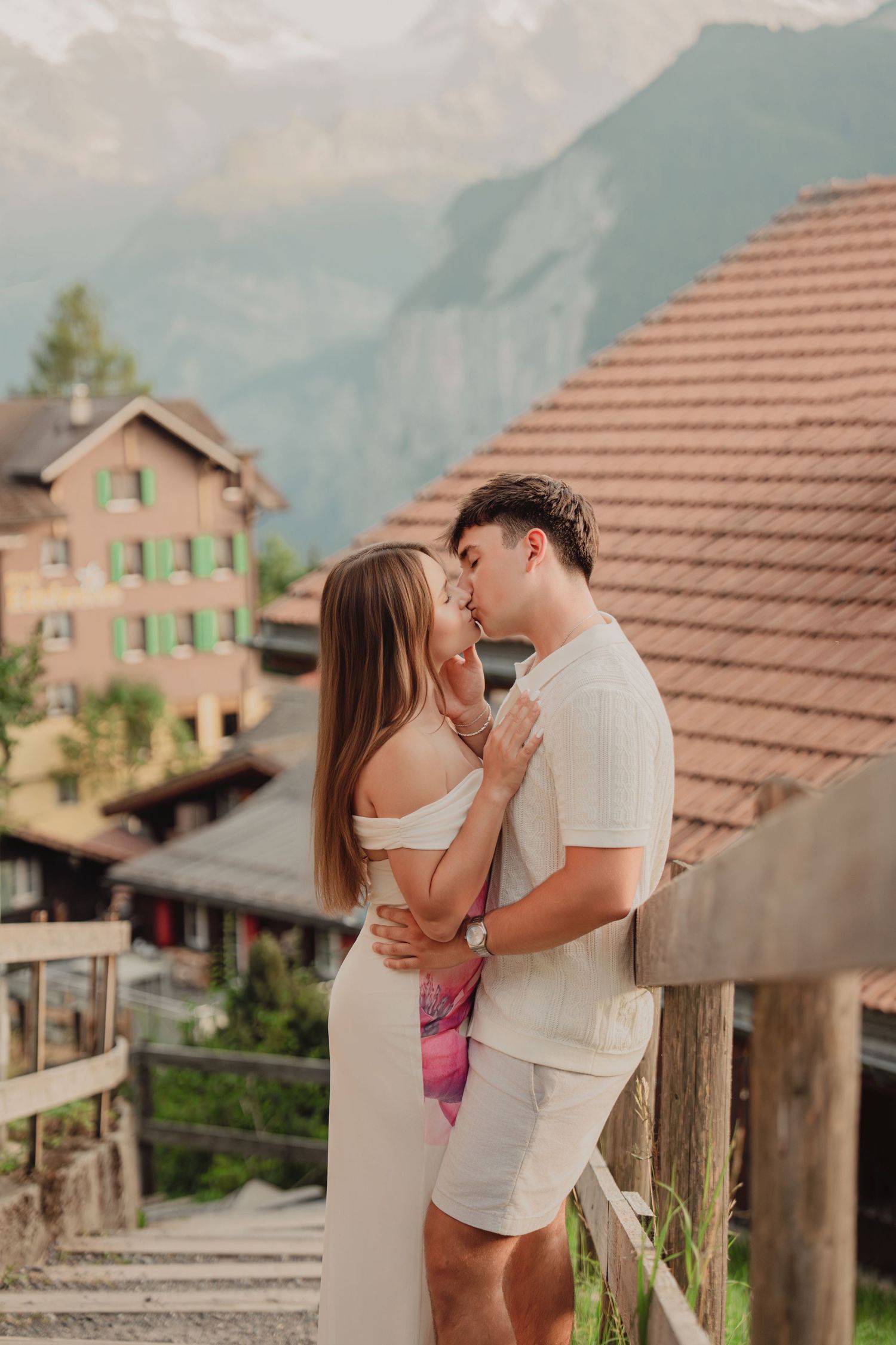 Couple sharing a kiss by wooden fence with Swiss alpine chalets and mountains behind them.