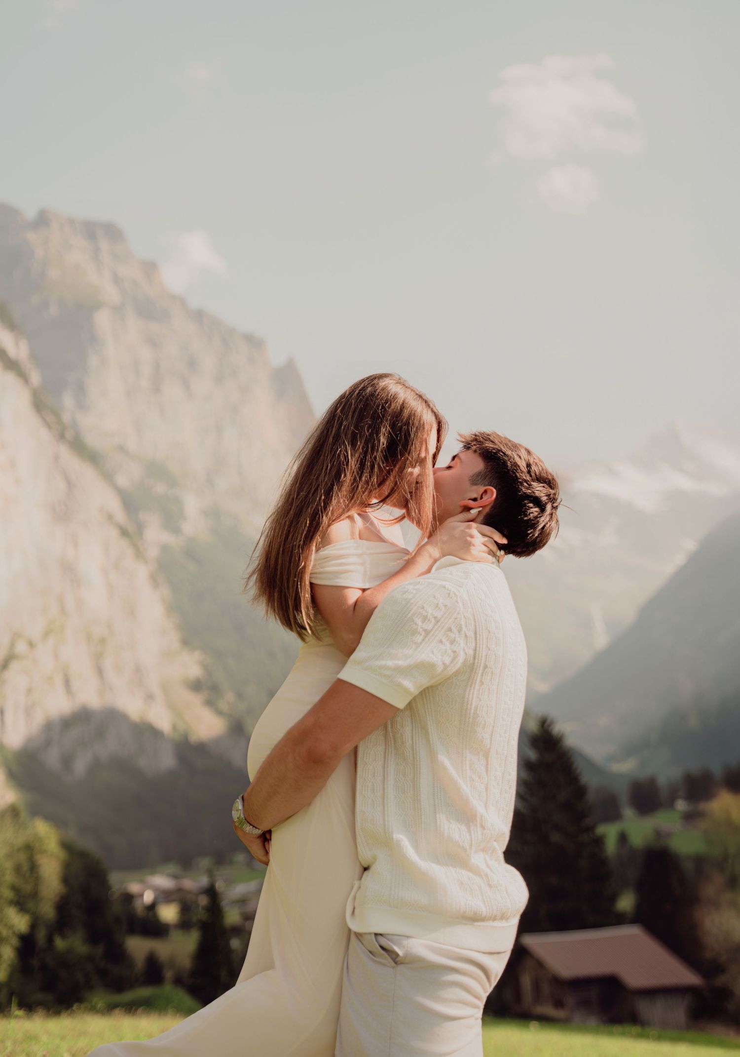 Romantic couple kissing in Swiss Alps meadow with dramatic mountain cliffs and valley below.