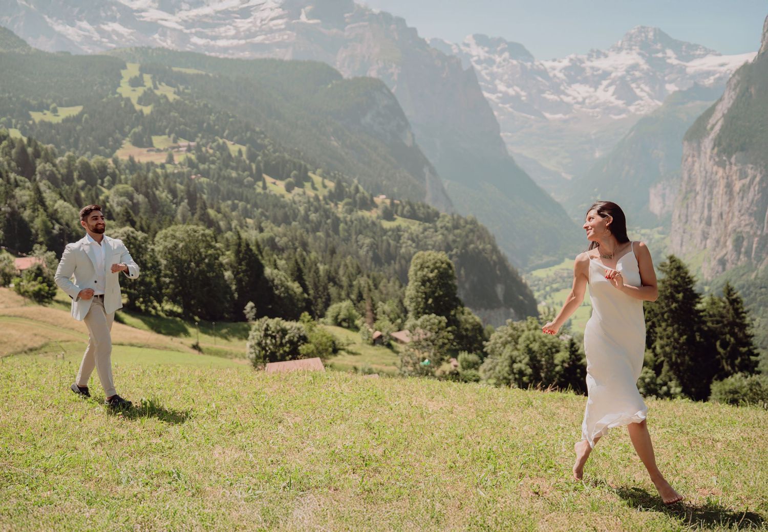 Couple playfully running across a lush green hillside with breathtaking Swiss Alps valley in background.