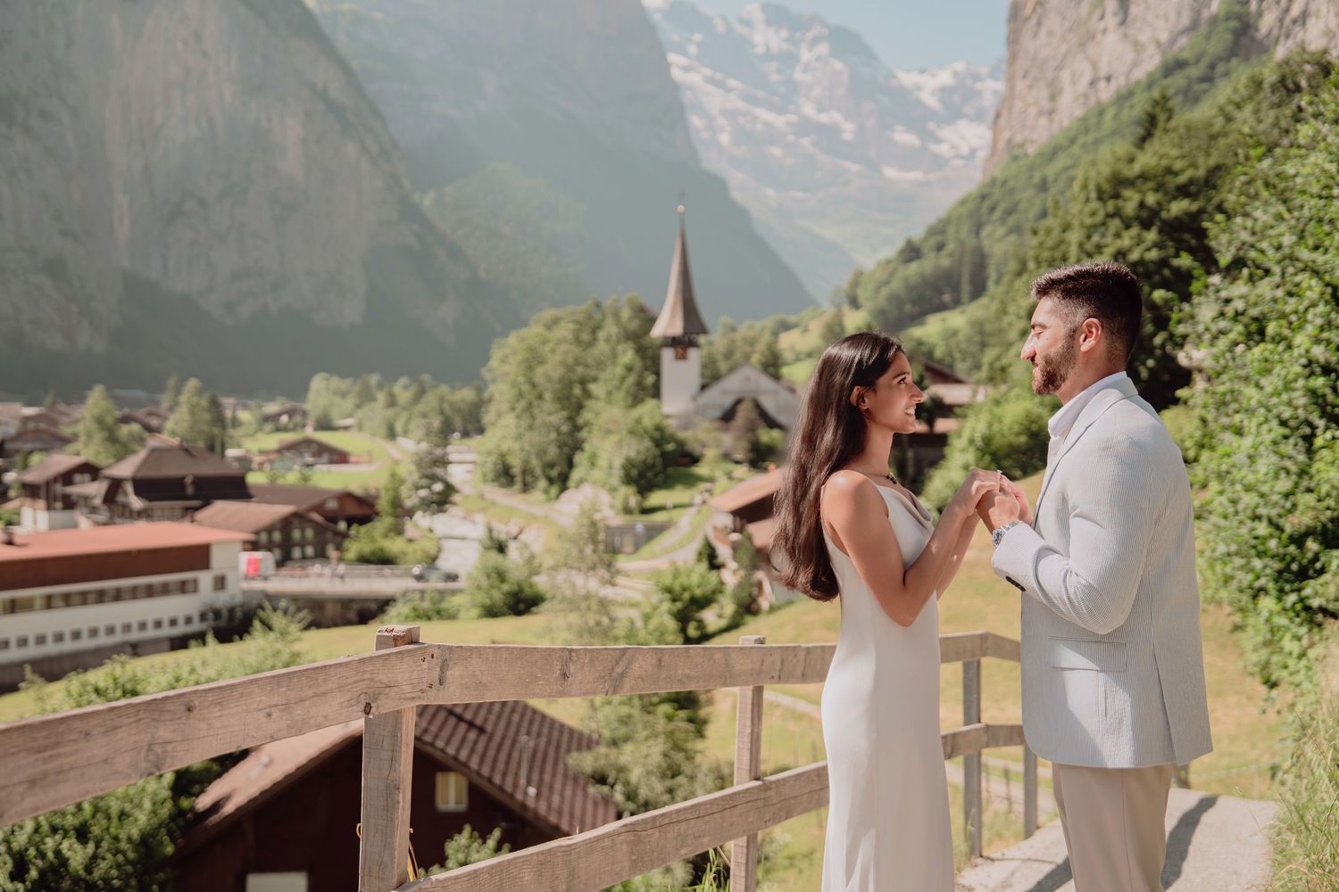 Couple shares a romantic moment on a wooden balcony overlooking a stunning Swiss Alpine village and mountains.