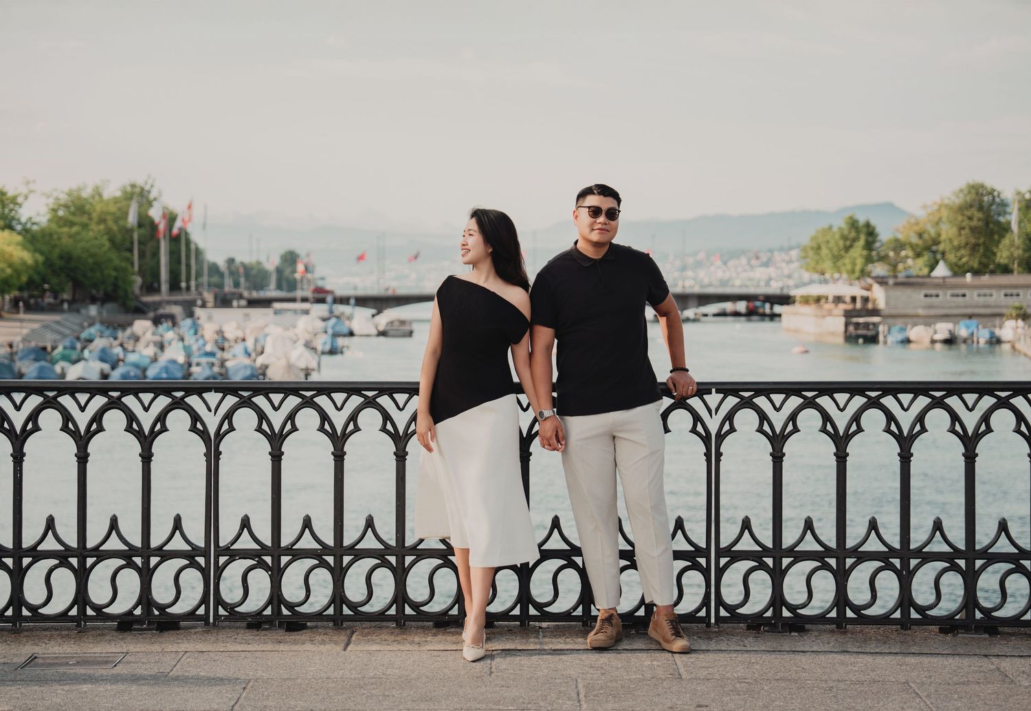 Couple holding hands back-to-back by ornate railing with Zurich lake and mountains backdrop.