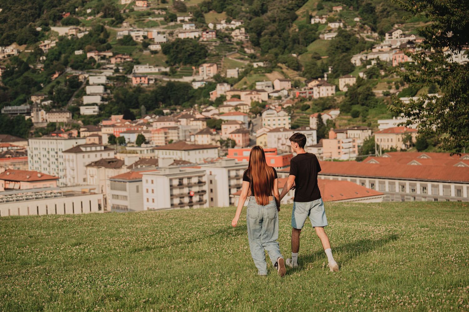 A couple walks hand-in-hand up a grassy hill overlooking a picturesque Italian hillside town.