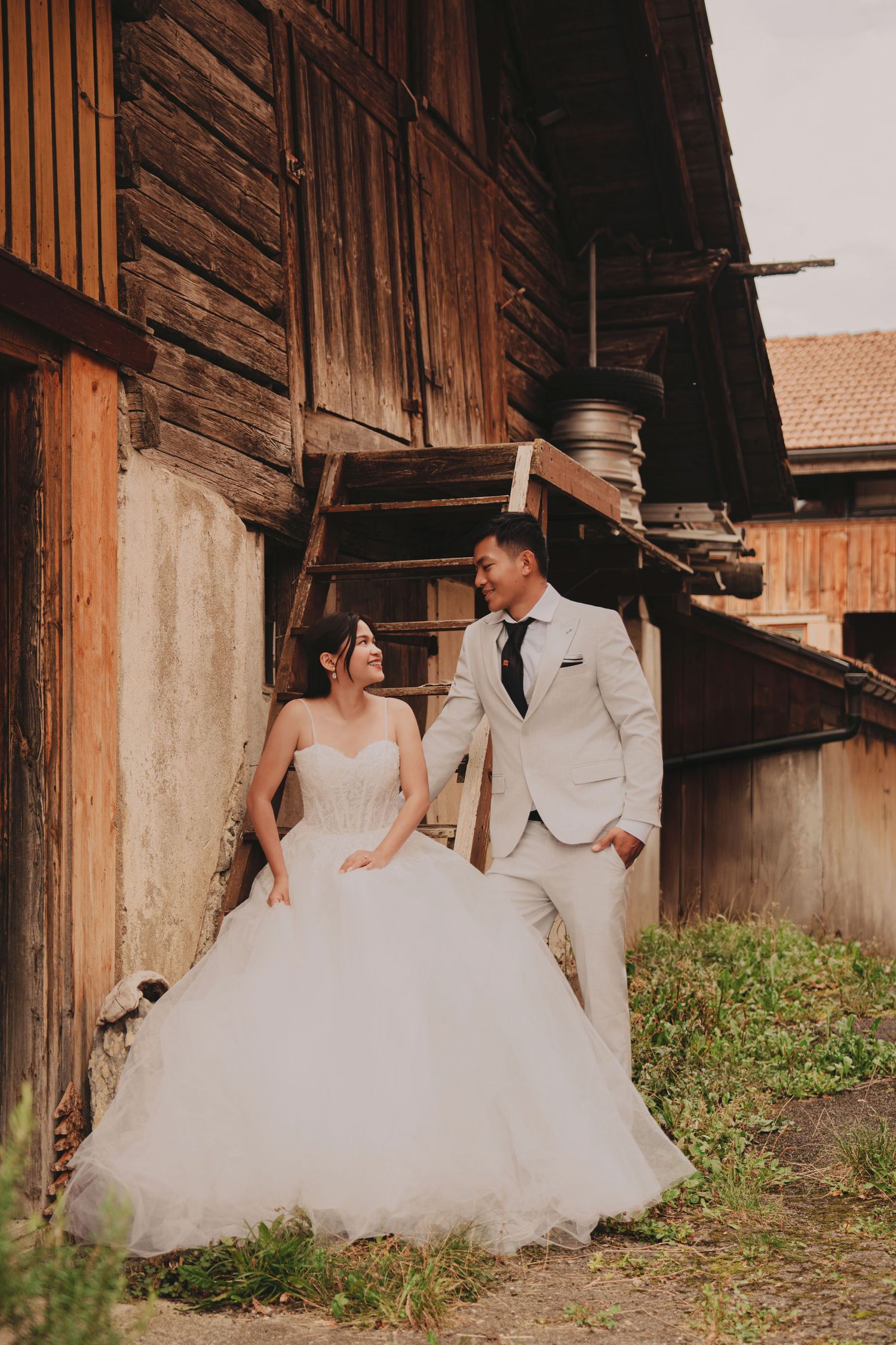 Bride and groom share a loving glance beside weathered wooden barn stairs in wedding attire.