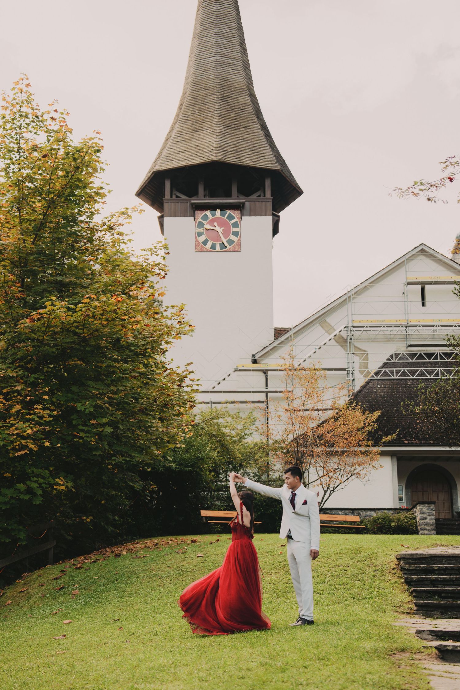 Couple dancing in red gown and white suit before Swiss church with ornate clock tower.