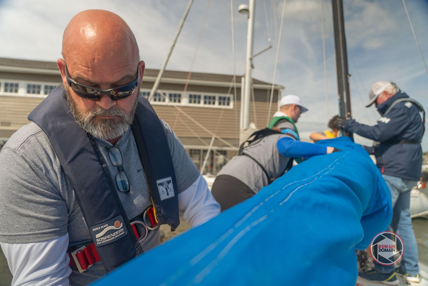 A group of people working together to fold and secure a large blue tarp or sail outdoors.