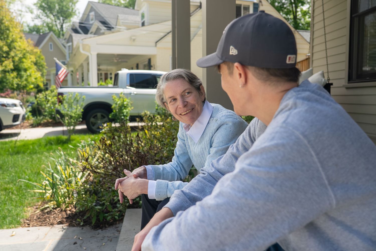 Two people sit on front porch steps, smiling and chatting in a sunny residential neighborhood with green landscaping.