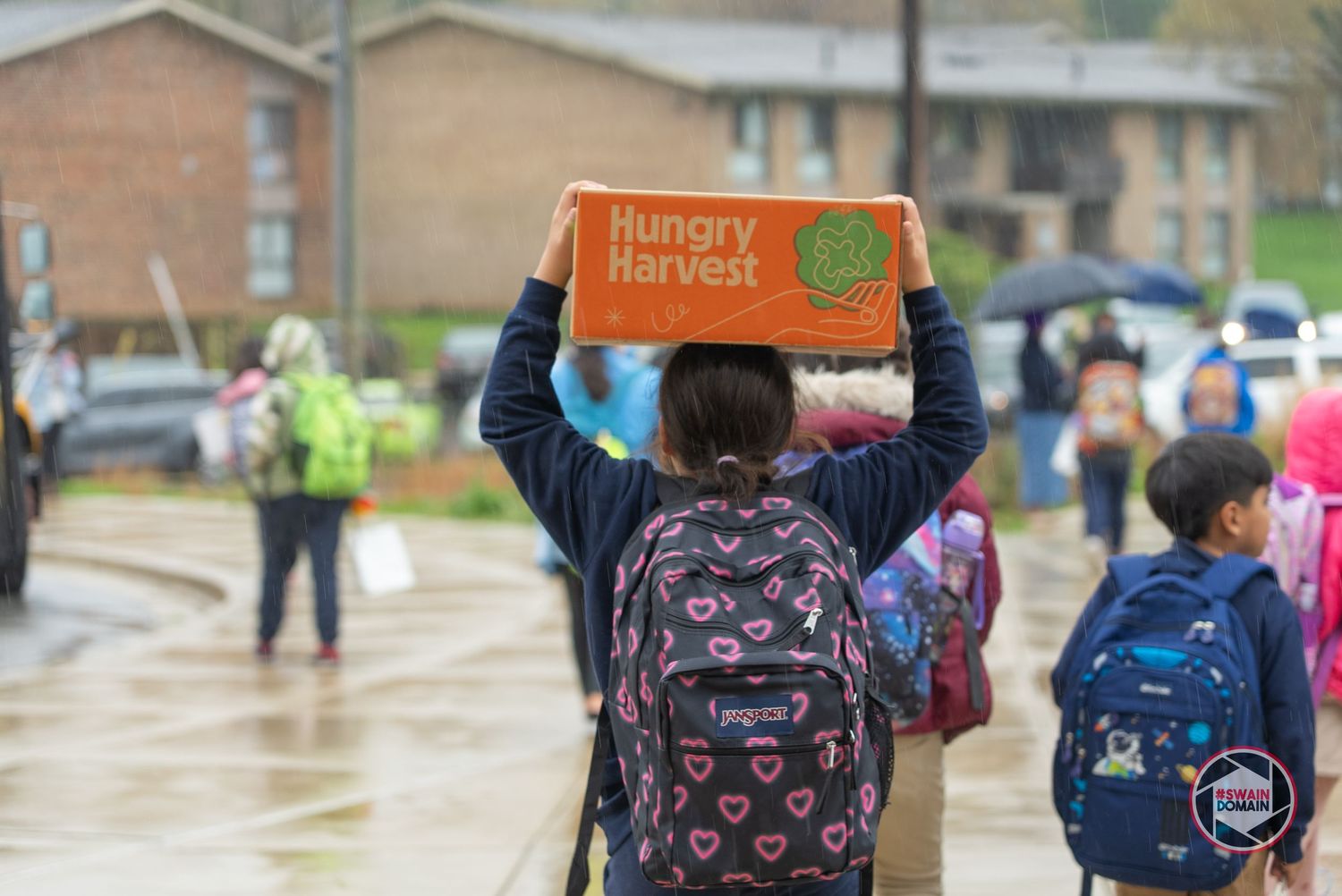 Students with backpacks carry orange boxes while walking outside a building.