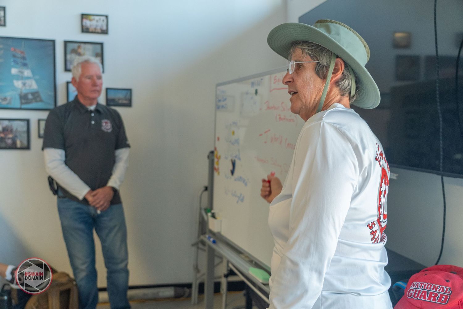 A person in a white jacket and baseball cap stands in front of whiteboards during what appears to be a presentation.