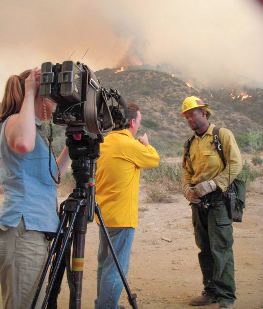 Camera crew filming outdoors in rugged terrain with professional equipment and yellow safety gear.