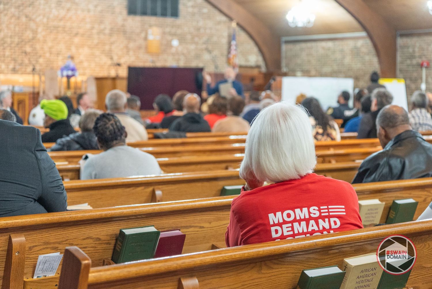 Woman in red Moms Demand Action shirt sits in church pew during a community meeting with audience members.
