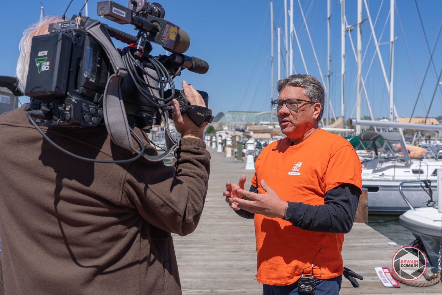 A person in an orange jacket being interviewed by a camera crew at a marina with boats in the background.