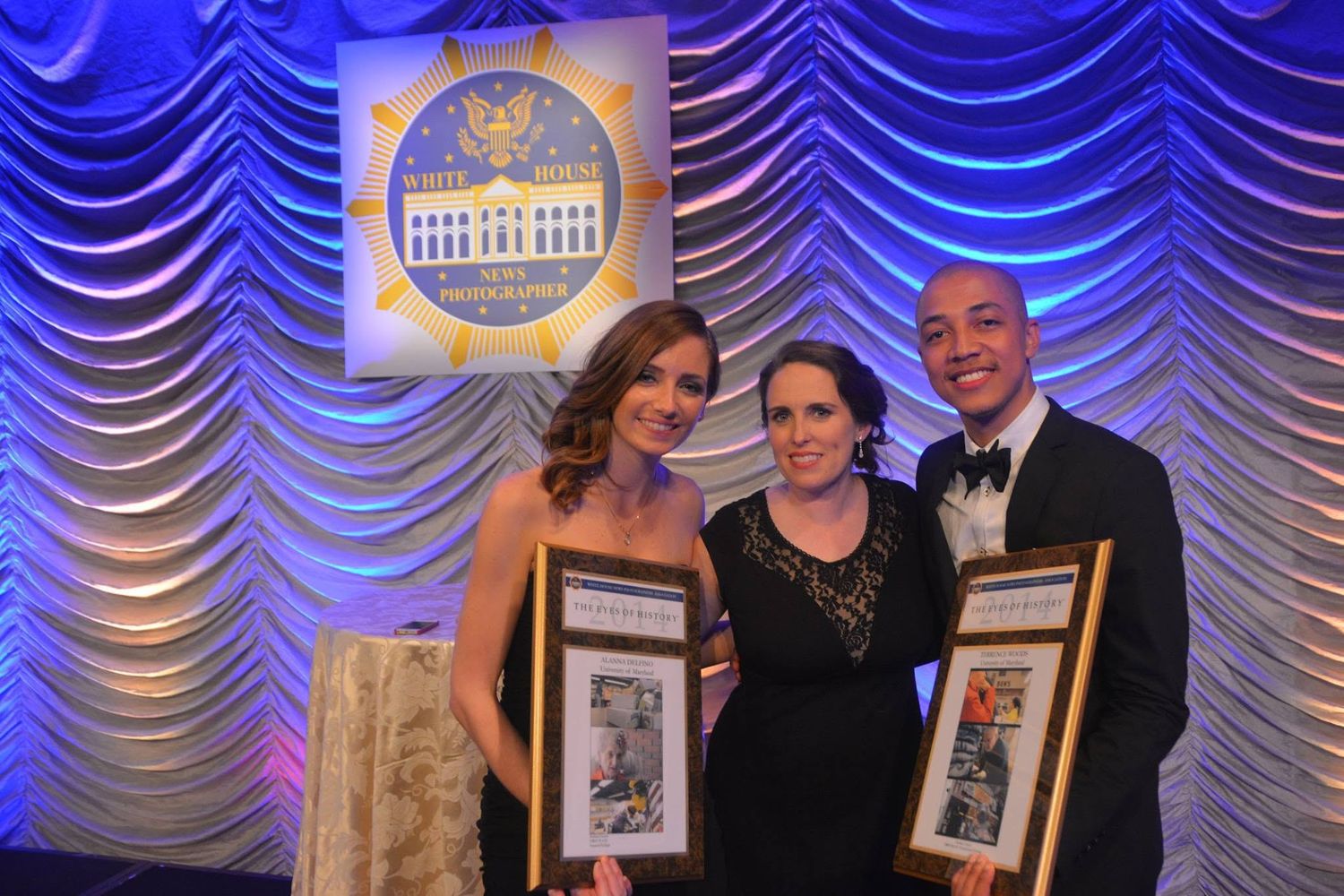 Three people posing with framed awards at a formal gala event with blue draped backdrop.