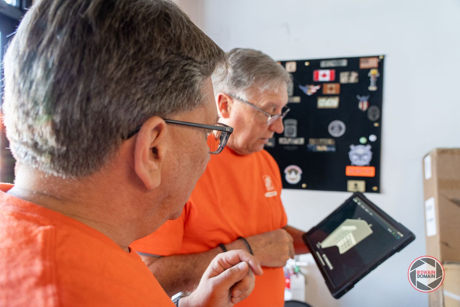 People in orange shirts examine and test electronic devices and equipment on a workbench.