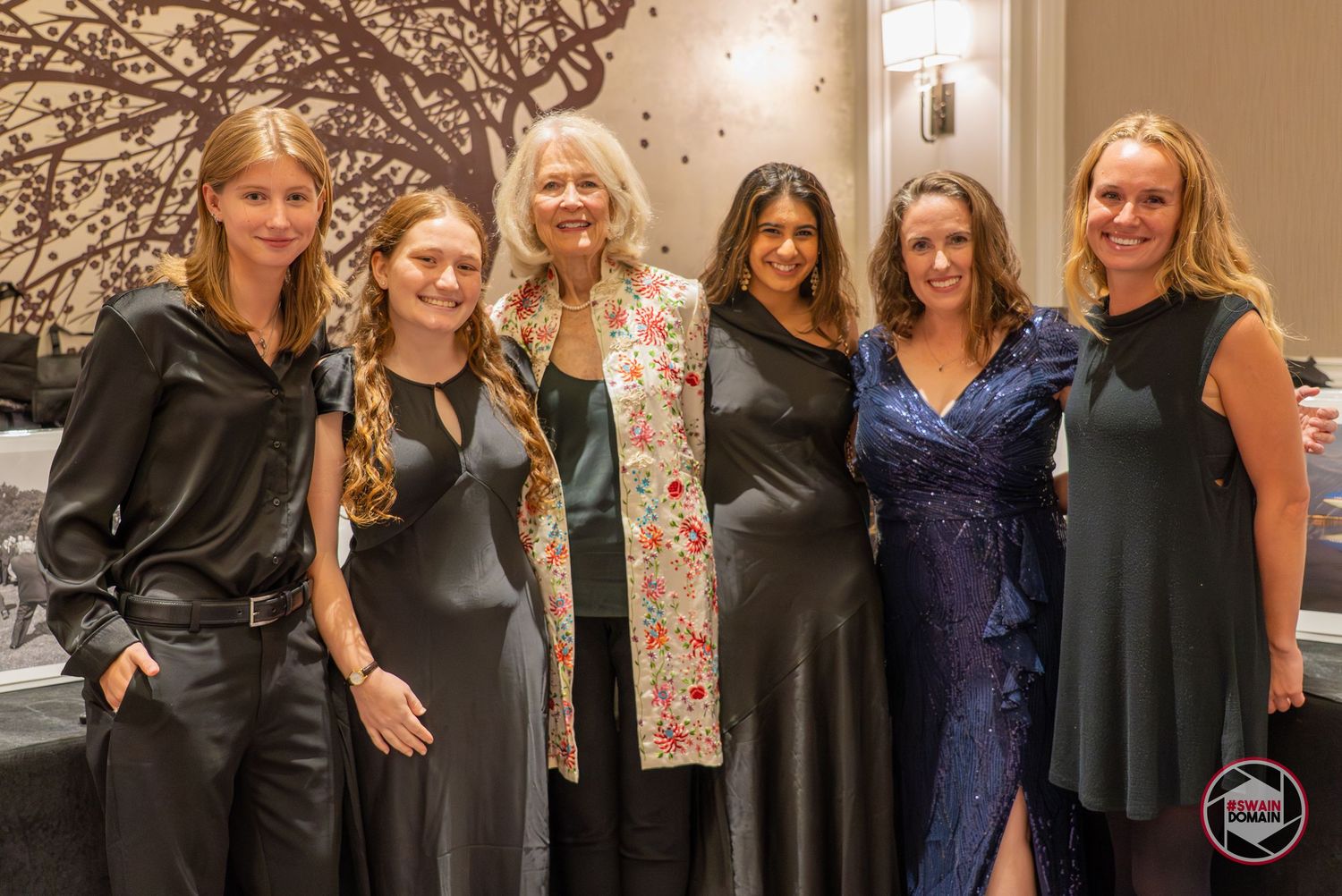 Six women posing together at a formal event, dressed in elegant attire, smiling for a group photo.
