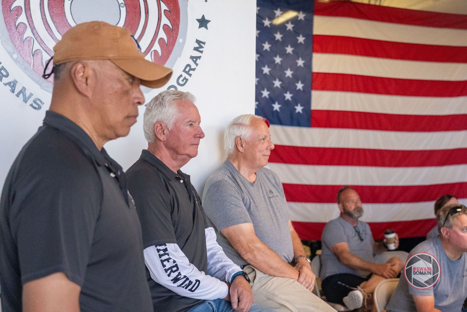 A row of people seated at tables with American flags displayed prominently on the wall behind them.
