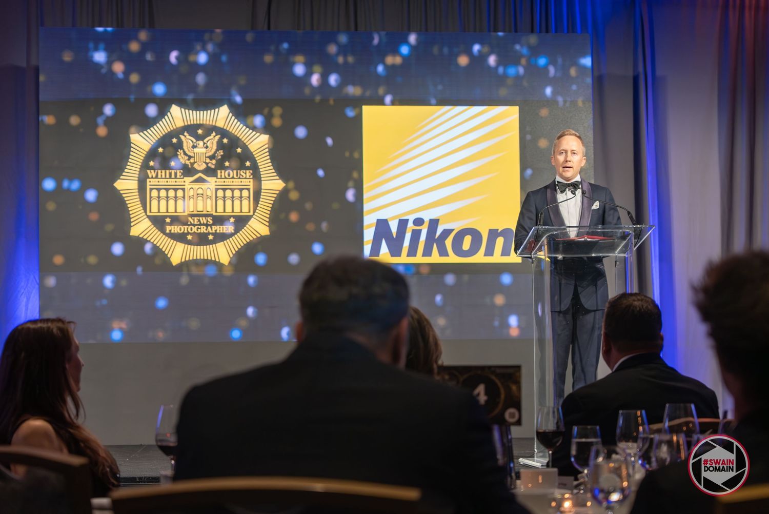 A speaker presents at a Nikon-sponsored awards gala with a decorative badge logo projected on screen behind him.