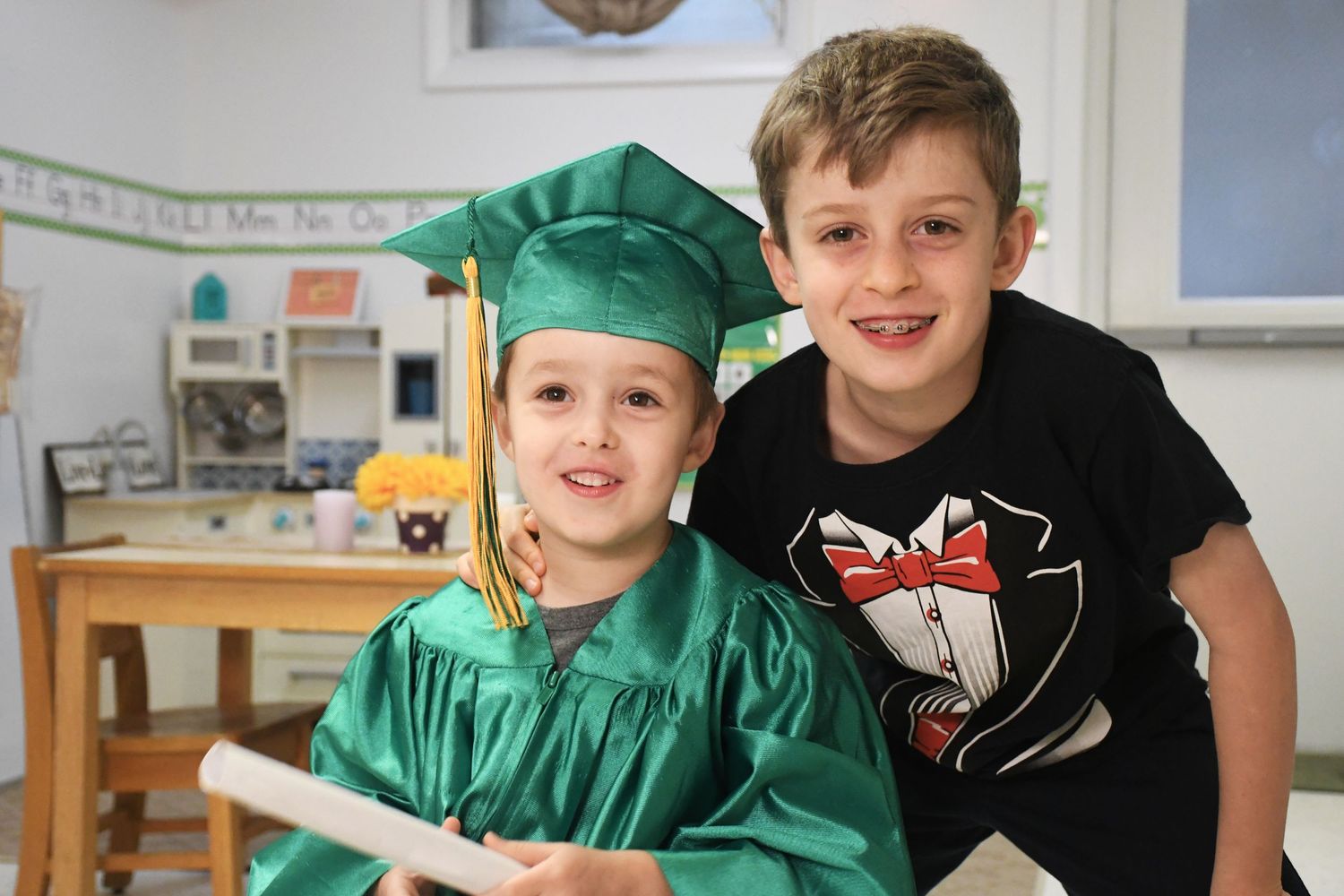 Young students wearing green graduation caps and gowns smile during a classroom celebration.