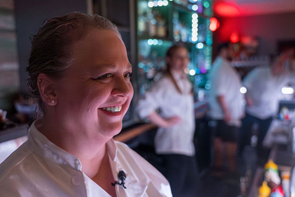 A person in chef whites smiles in a dimly lit restaurant kitchen.
