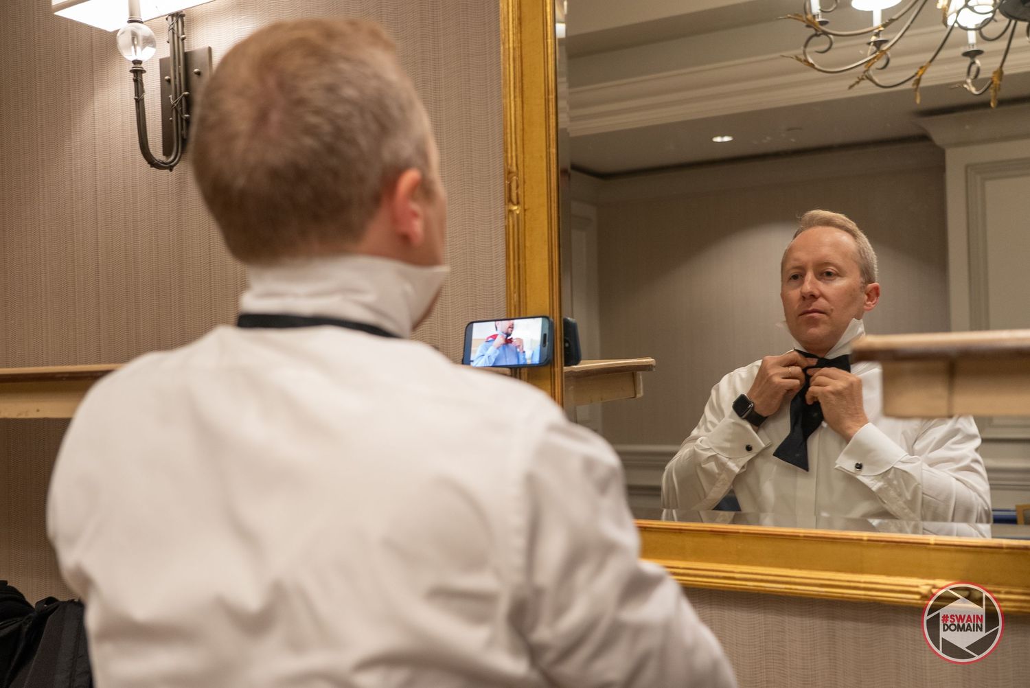 Man adjusting bow tie while looking in ornate gold mirror, with phone propped up reflecting his reflection.