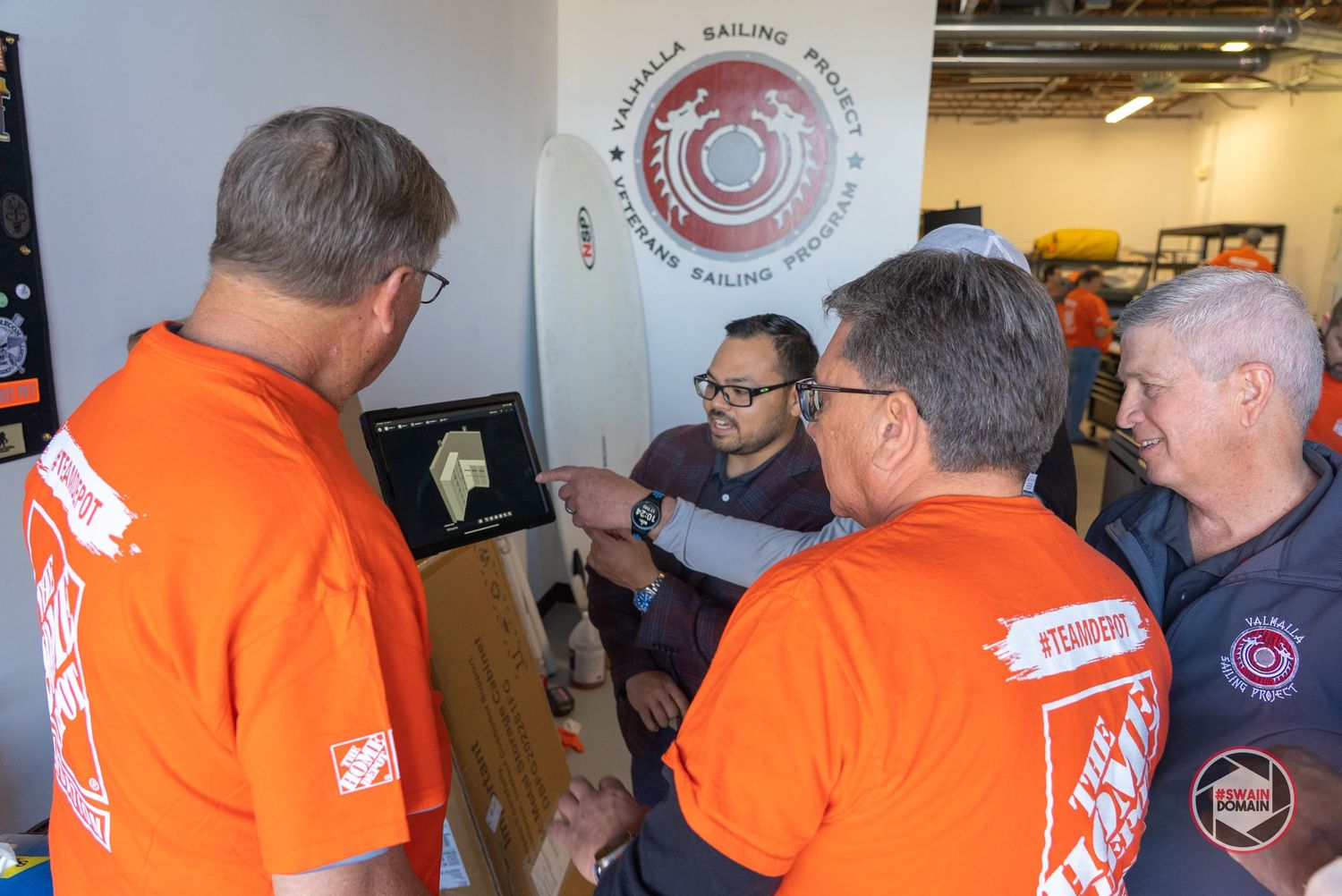 People in bright orange jackets gather at an indoor event with circular logo signage visible on the wall.
