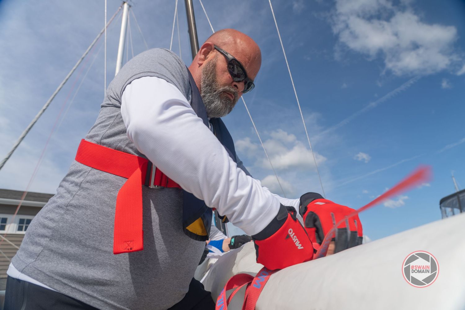A person in a gray jacket and sunglasses attaches red safety lines to equipment on a sunny day with blue skies.
