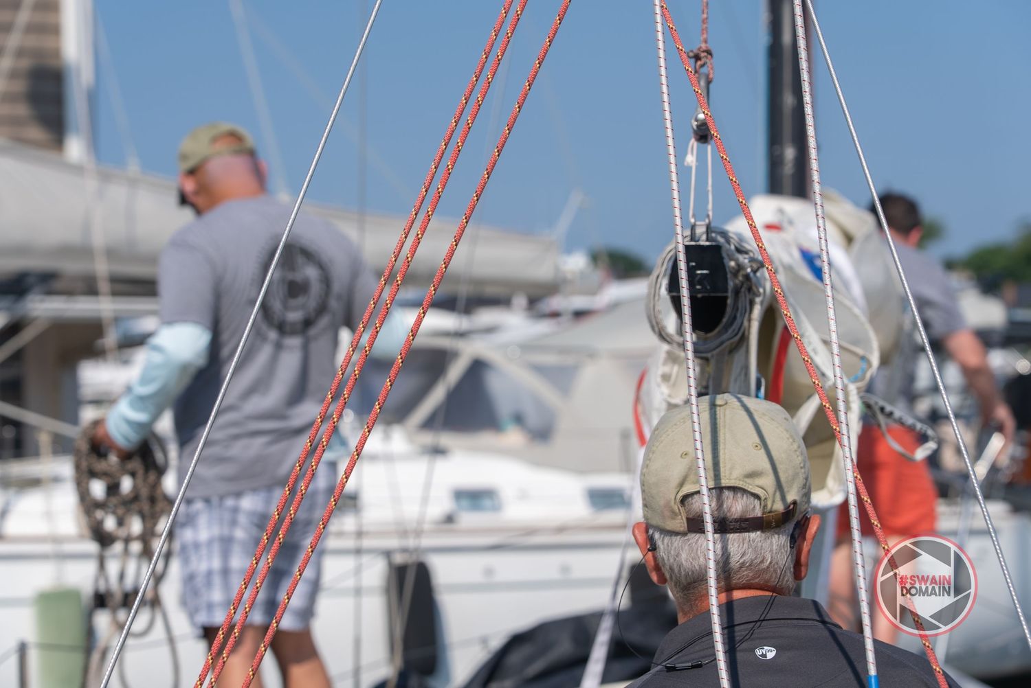 A sailboat mast with red and white rigging equipment against a bright blue sky and marina background.