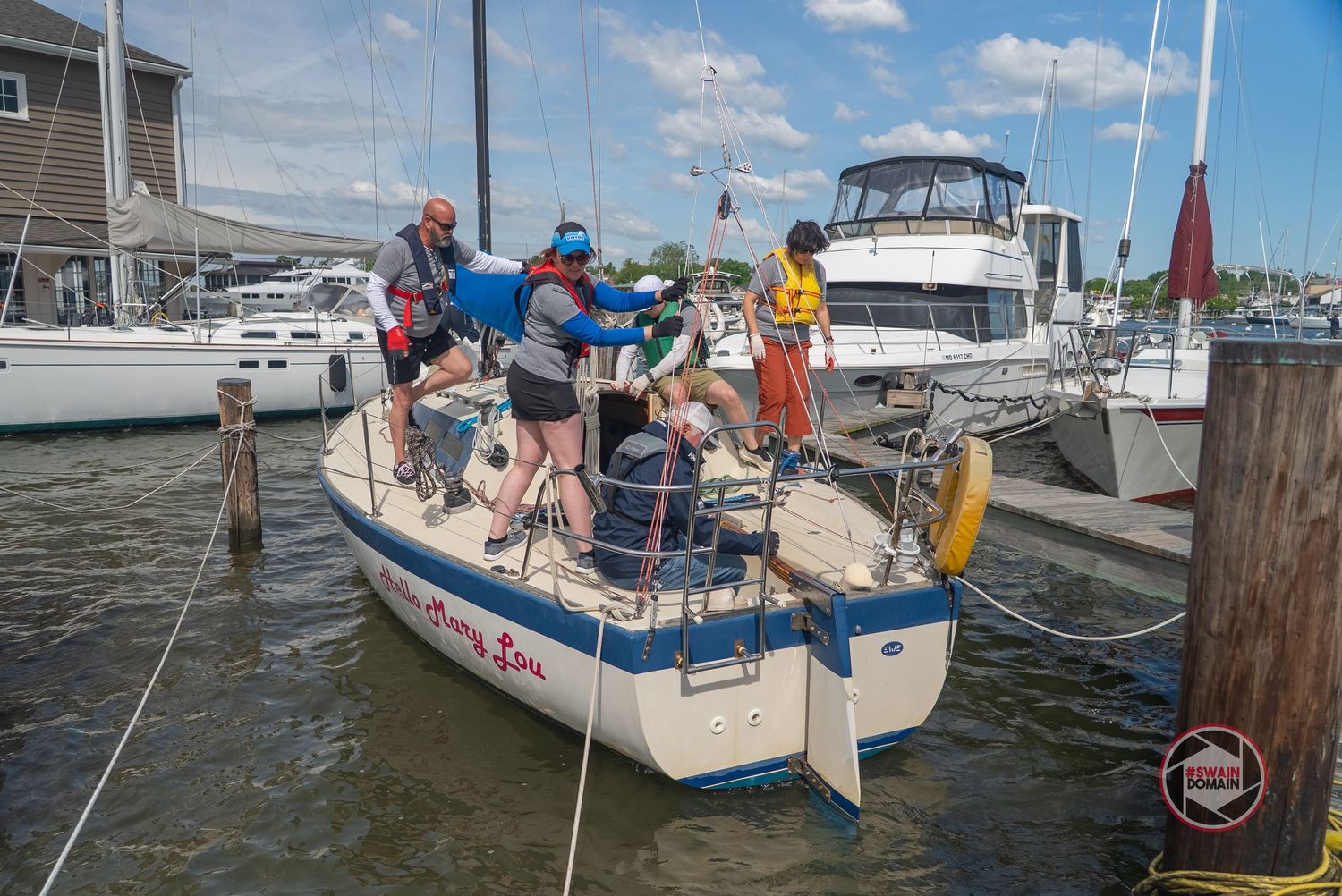 A group of people gather on a sailboat docked at a marina on a sunny day with other boats visible in the background.