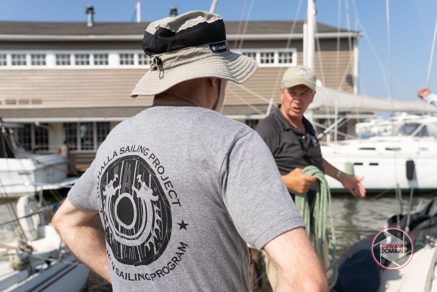 Two people conversing at a marina dock with boats and a yacht club building visible in the background.