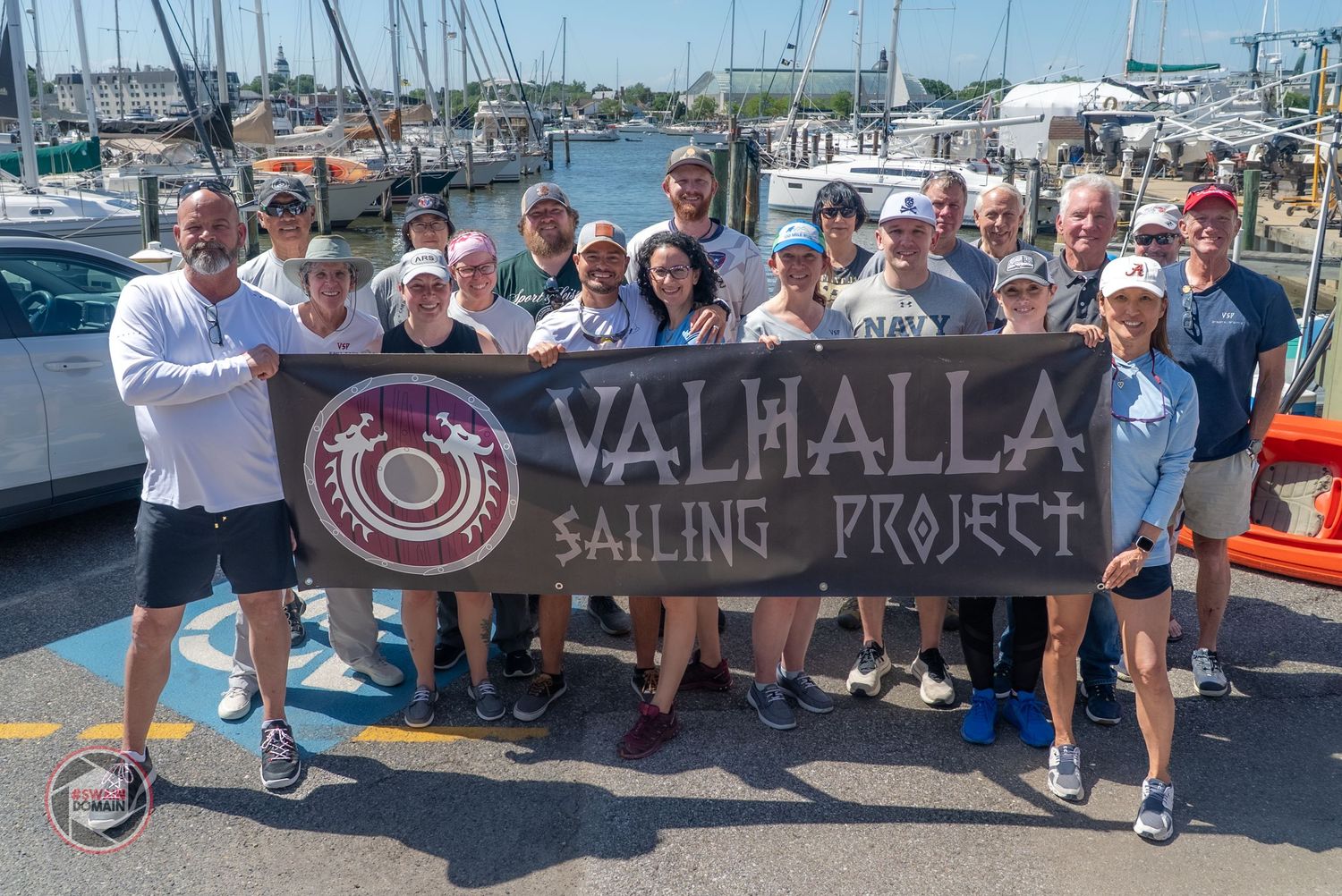 A group of people gather around boats and signs at a marina during a sunny day.