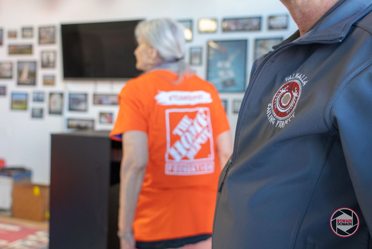 Two workers in contrasting orange and navy uniforms stand in an office setting with photos on the wall.