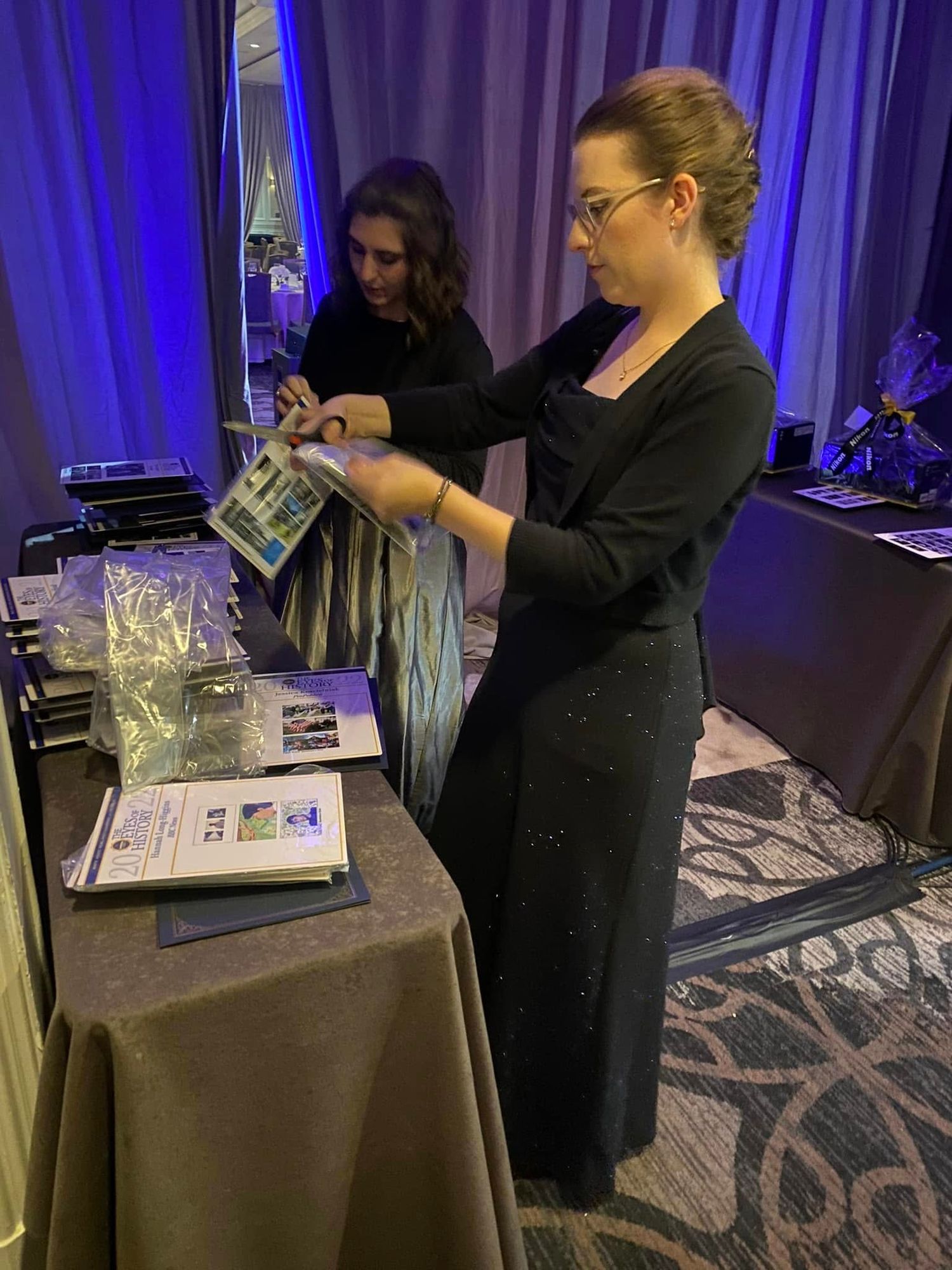 Two women in black formal attire organizing materials at event registration table in elegantly lit banquet hall.