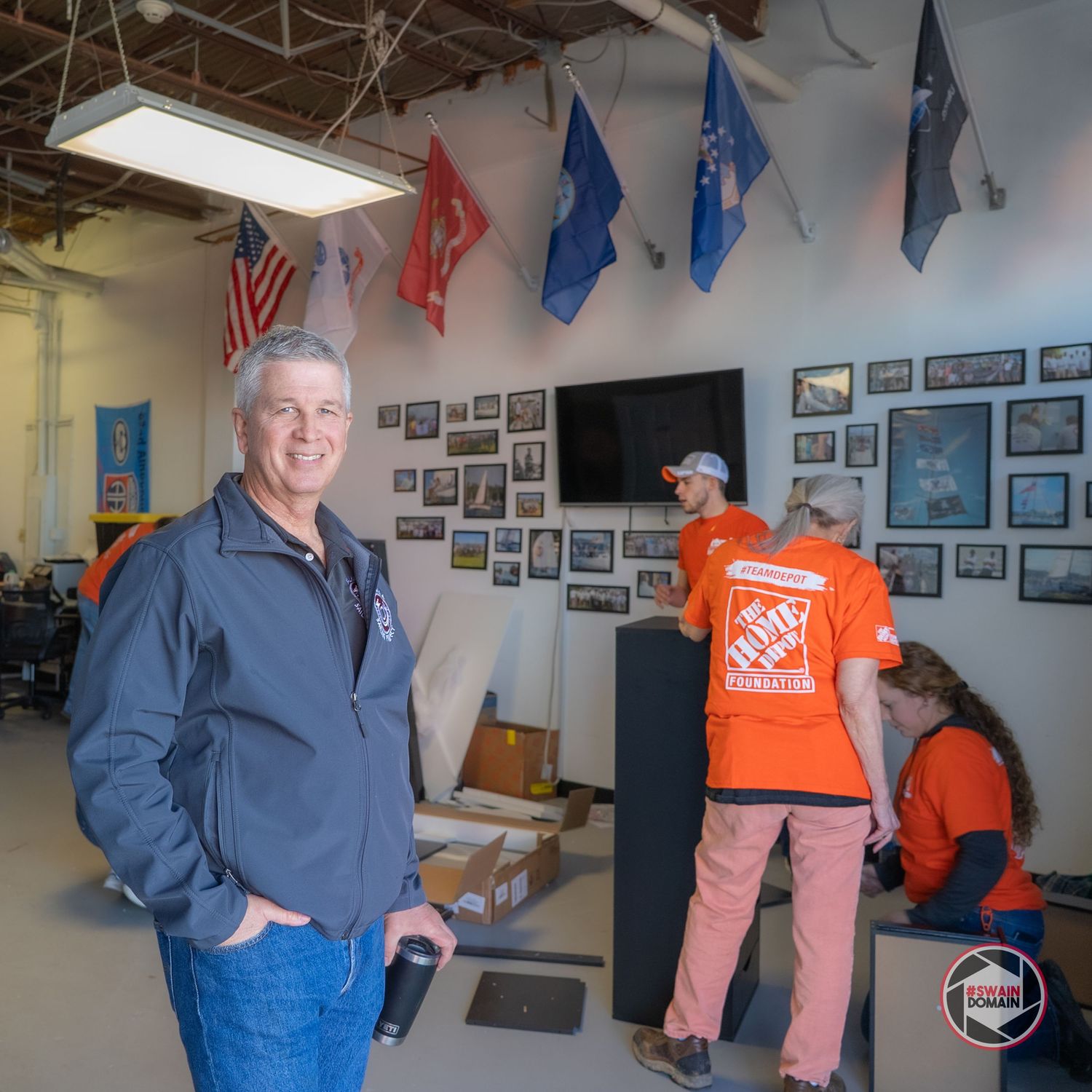 Workers in blue uniforms and orange Home Depot aprons gather in a room decorated with flags and photos on the walls.