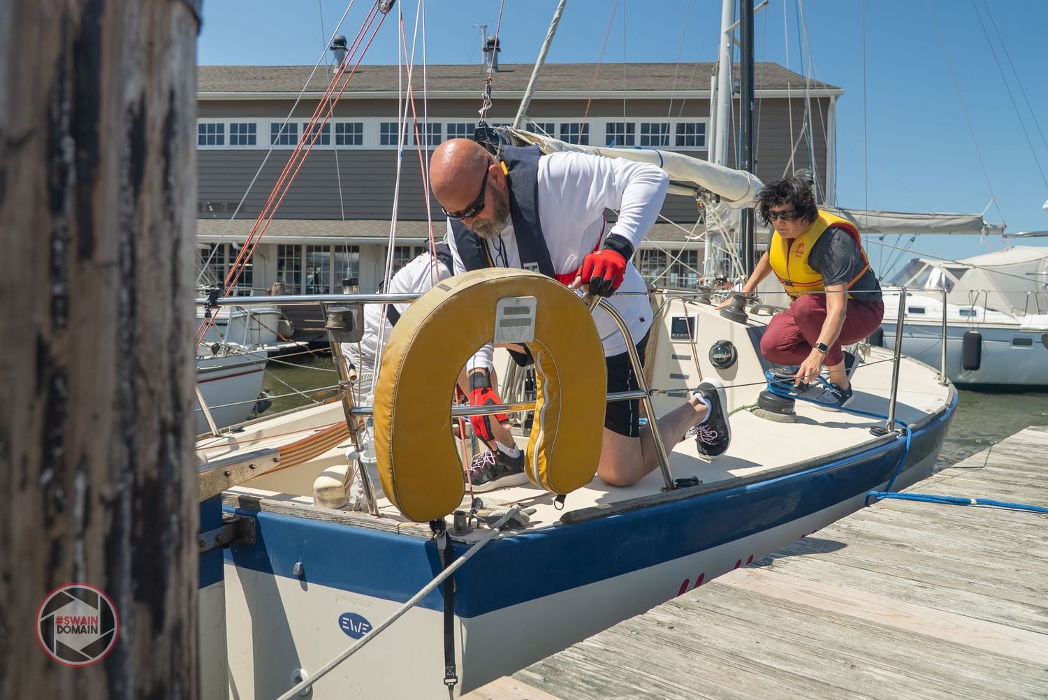 A series of sailboats docked at a marina with yellow equipment visible on the boats under sunny blue skies.