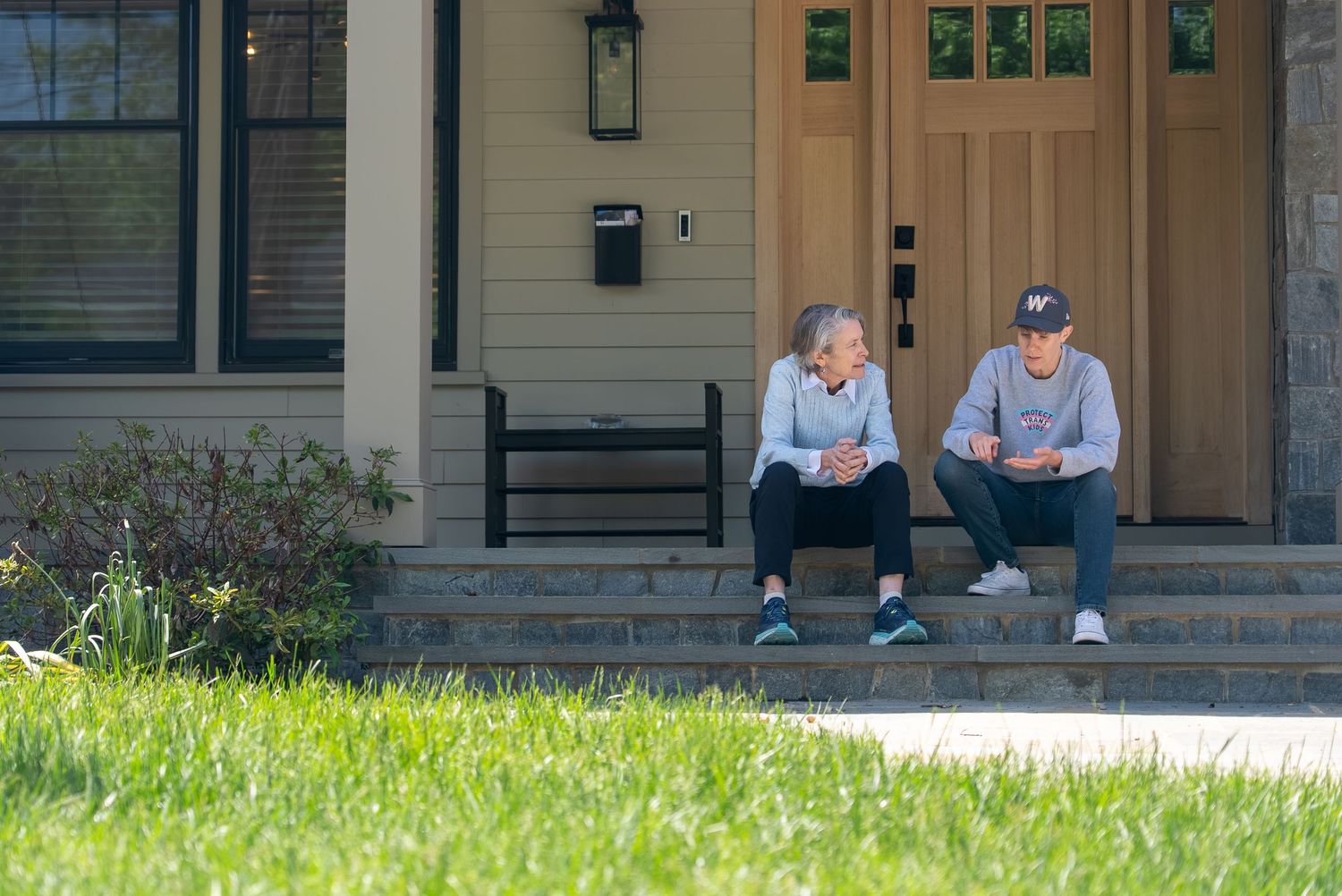 Two people sitting on front porch steps of a modern home, relaxing outdoors on a sunny day.