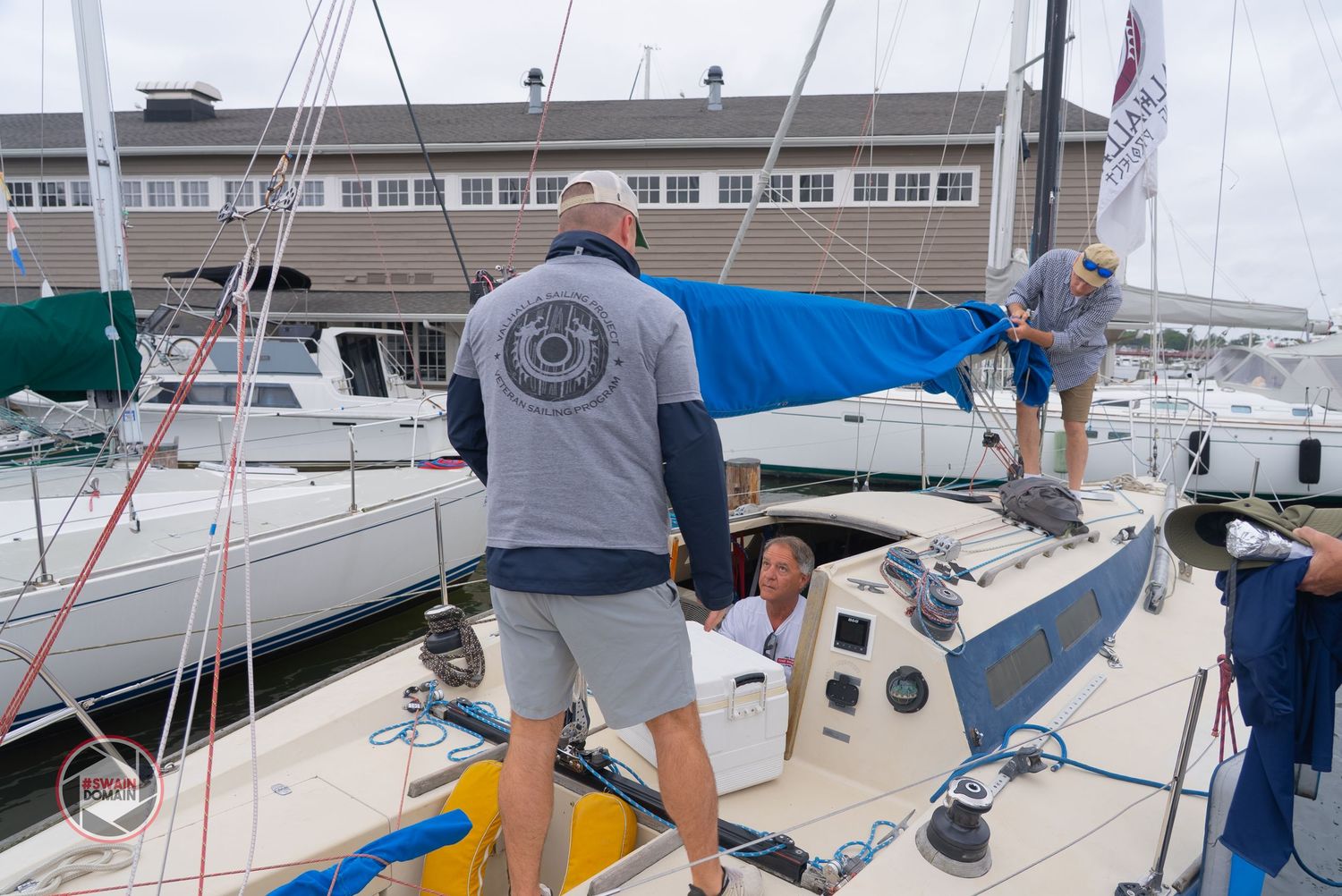 Crew members work on preparing a sailboat at a marina dock with boats and buildings in the background.