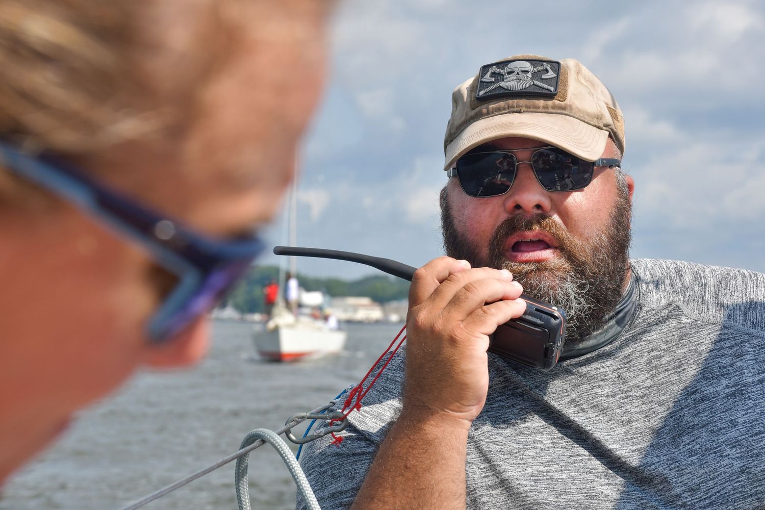 A bearded person in sunglasses and a baseball cap steers a boat while speaking into a radio on the water.