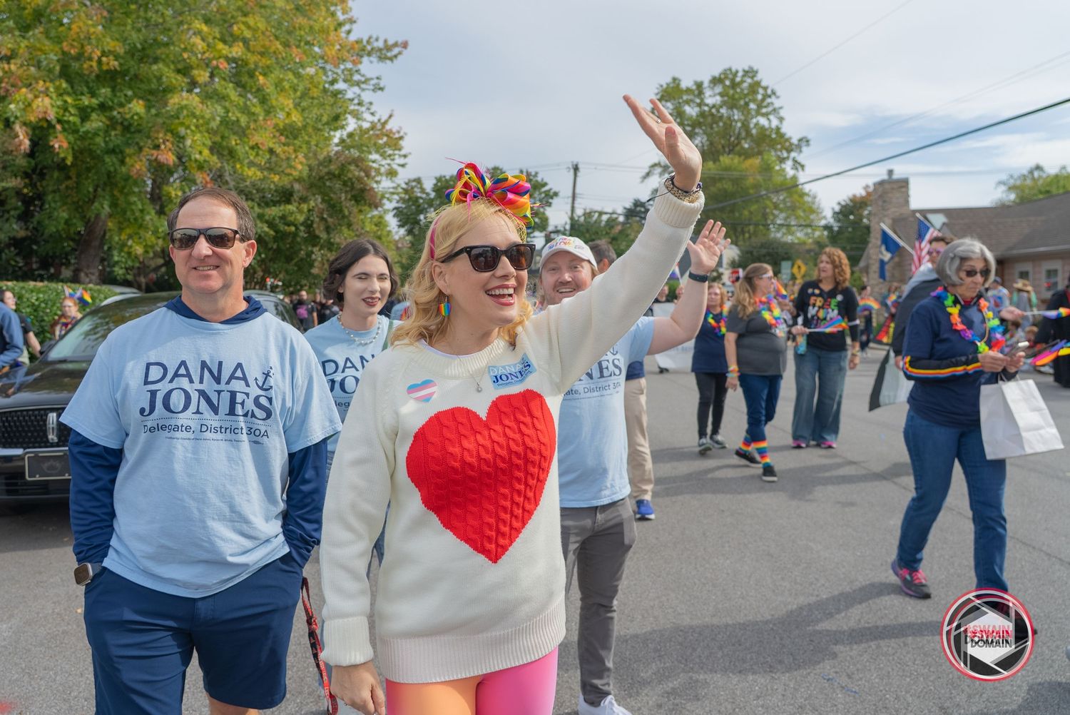 People marching in a parade, wearing Dana Jones campaign shirts, waving to crowds on a sunny day.