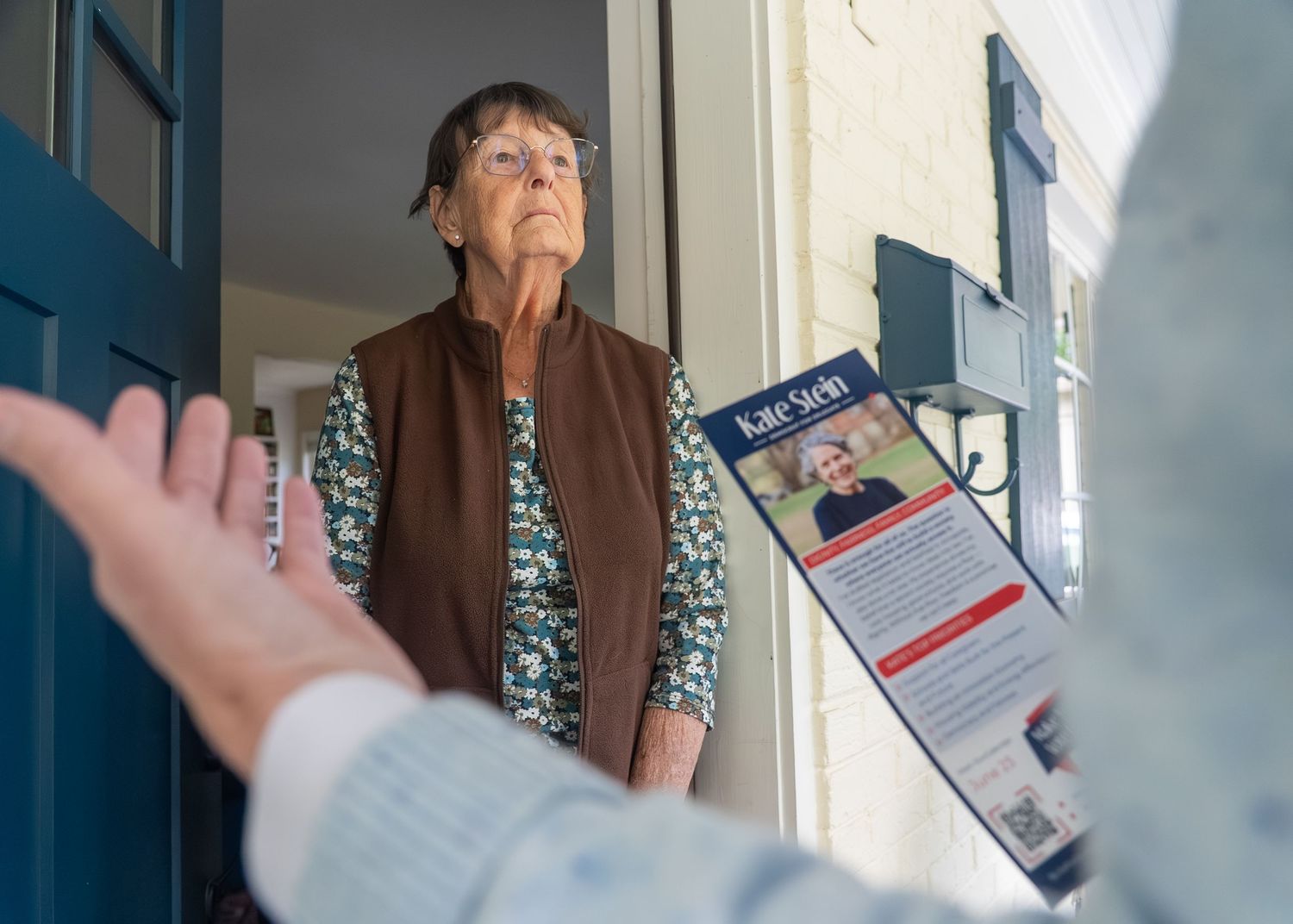 Elderly woman at front door receiving a campaign flyer from a canvasser during a political door-to-door outreach visit.