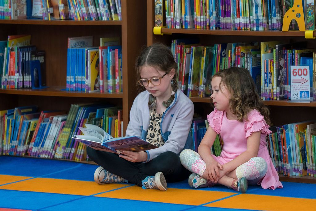 Students reading books together on the floor of a colorful library with bookshelves in the background.
