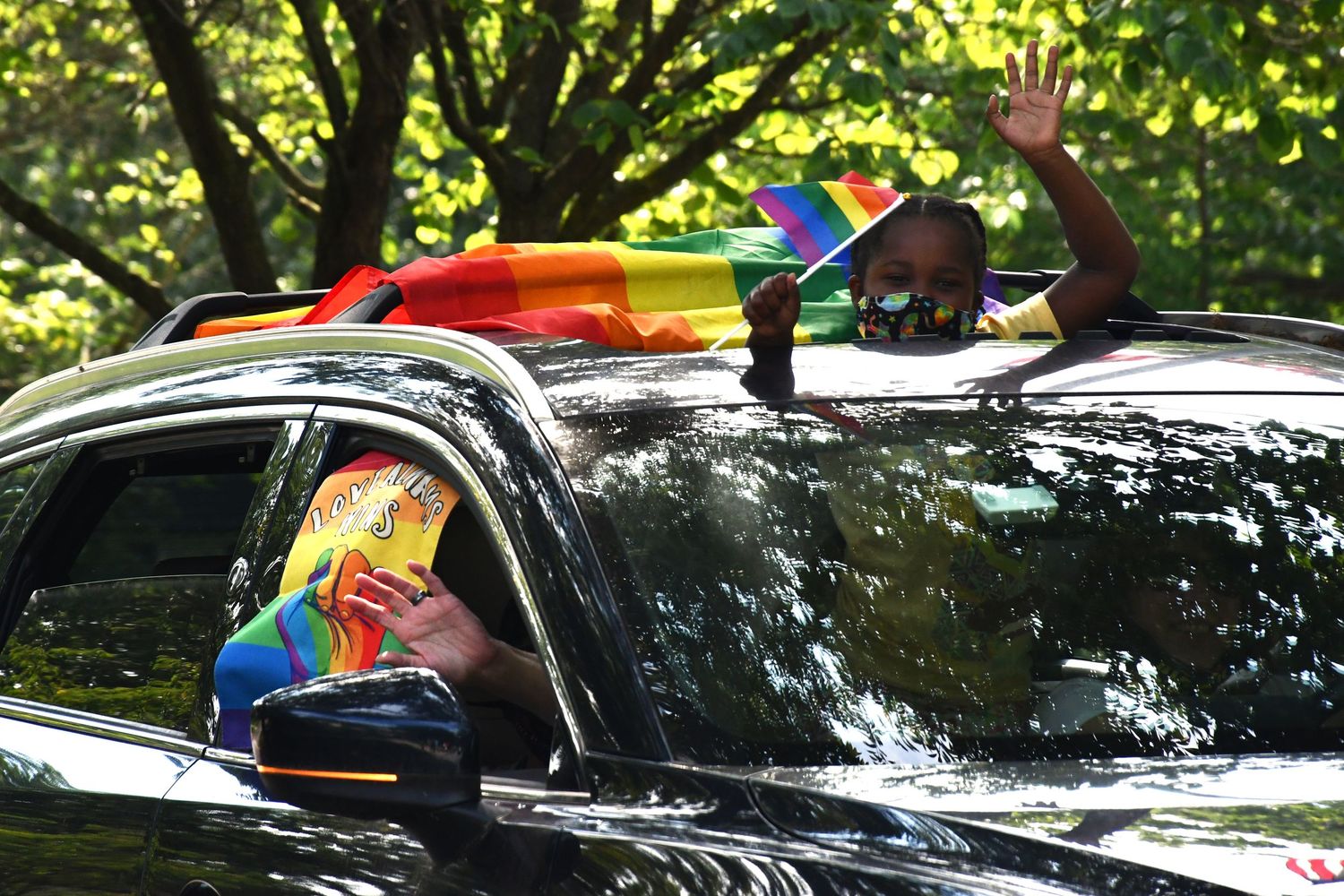 Cars line up in a sunny wooded area with rainbow flags visible.