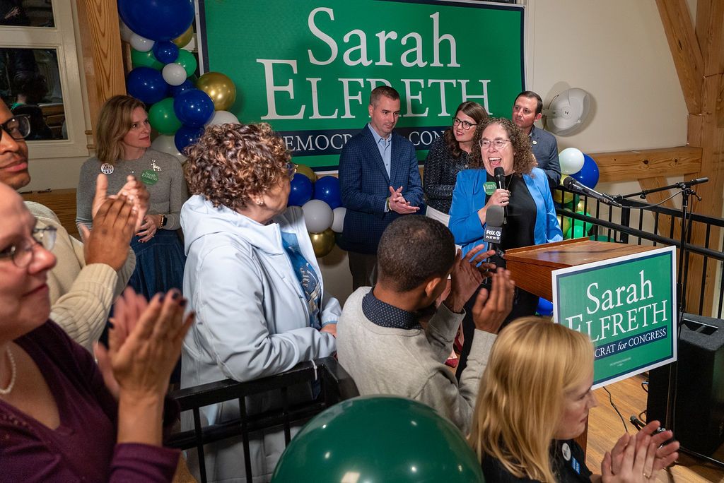 Political rally with green campaign signs and supporters gathered in indoor venue.