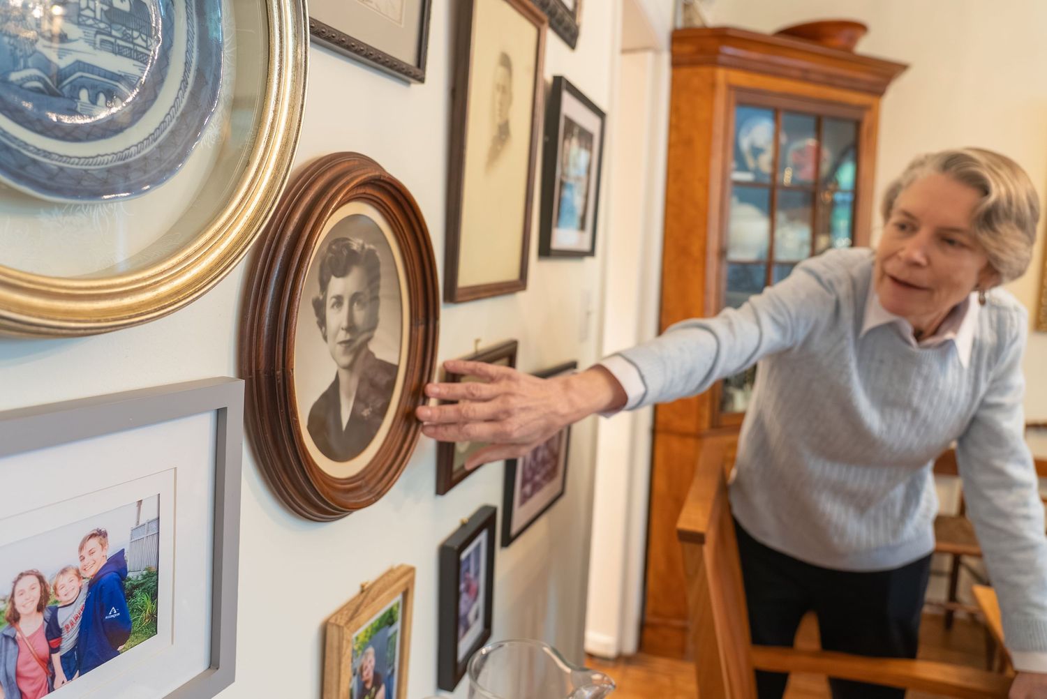 Elderly woman touching a framed vintage portrait on a wall gallery of family photos in a home interior.