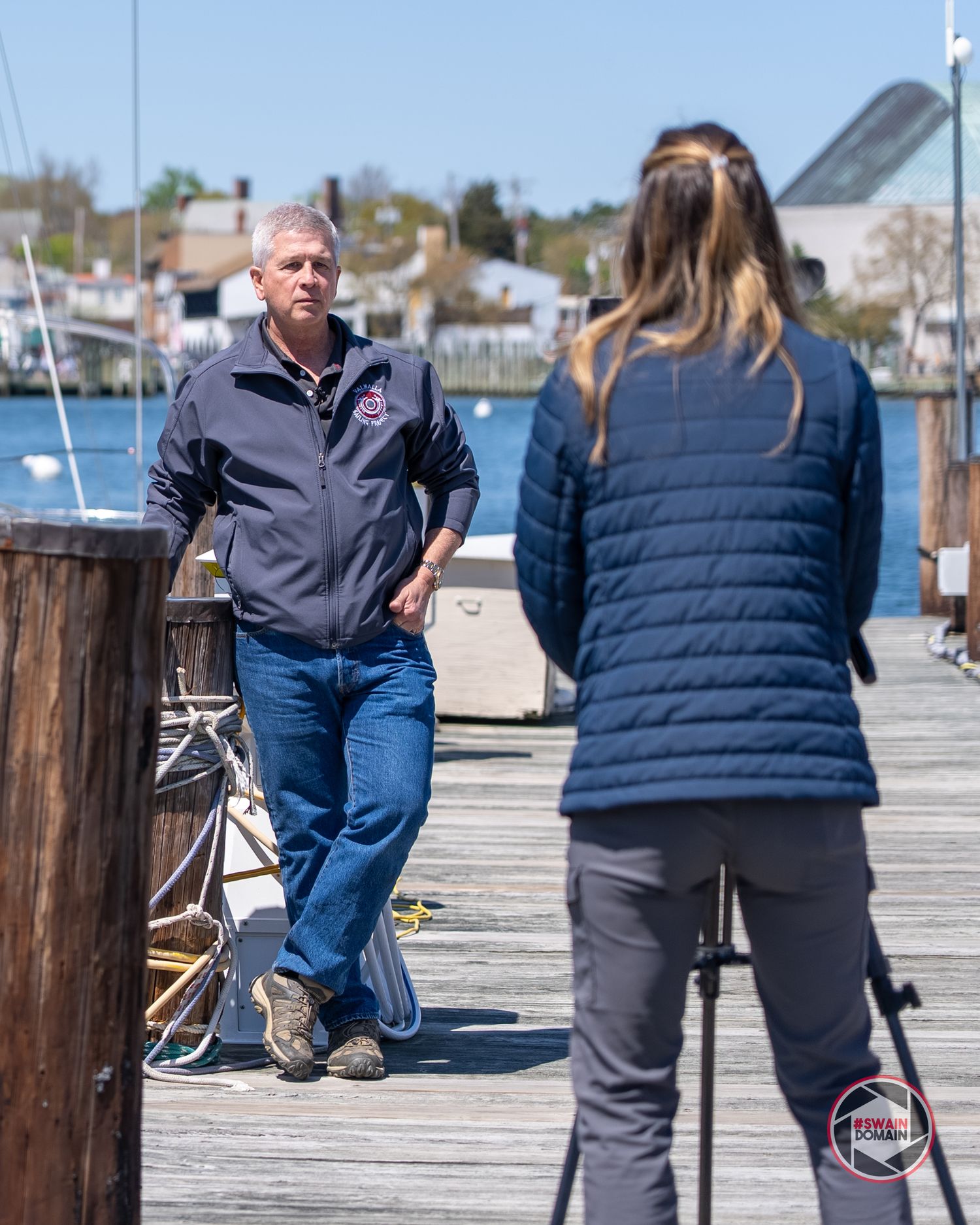 People in winter coats walking along a wooden dock by a marina with boats and water in the background.