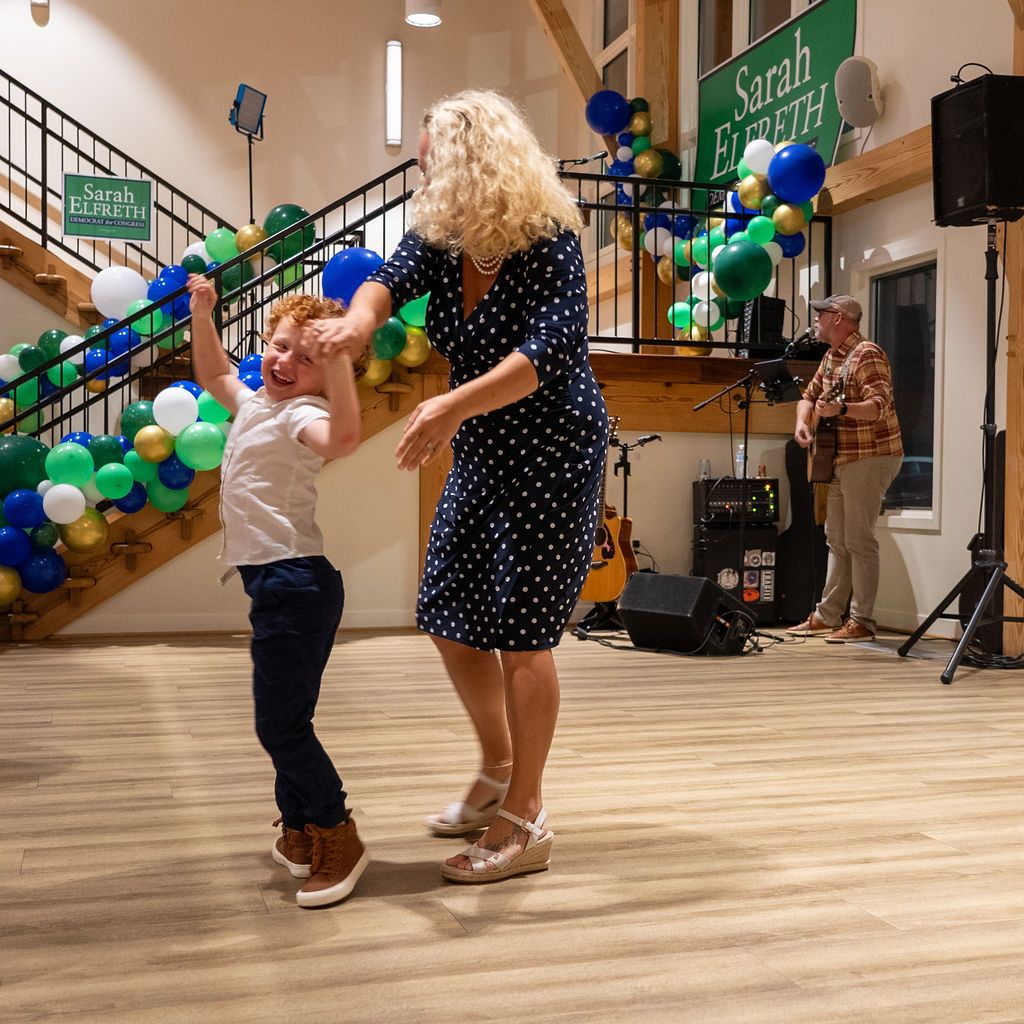People dancing and celebrating at a political campaign event with blue and green balloons.